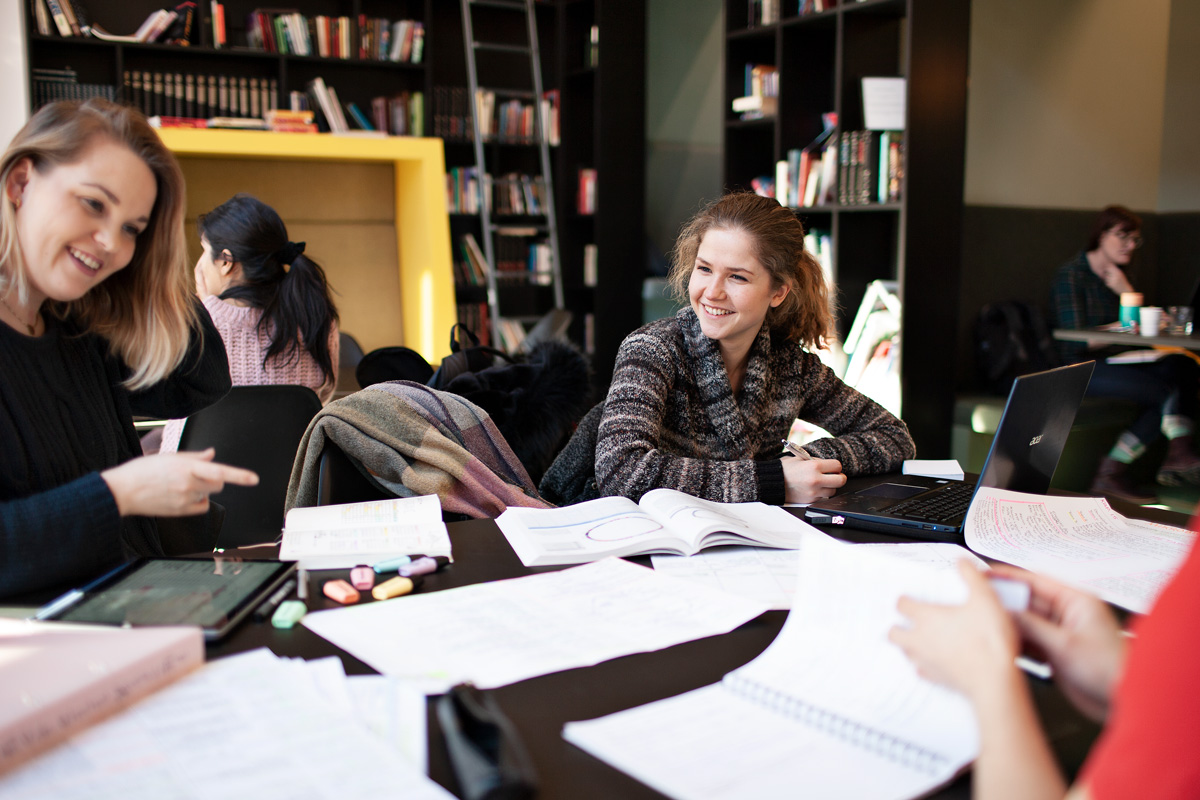 Tre studenter som sitter rundt bord i studiemiljø med bøker, PC og annet studiemateriell på bordet. Foto.