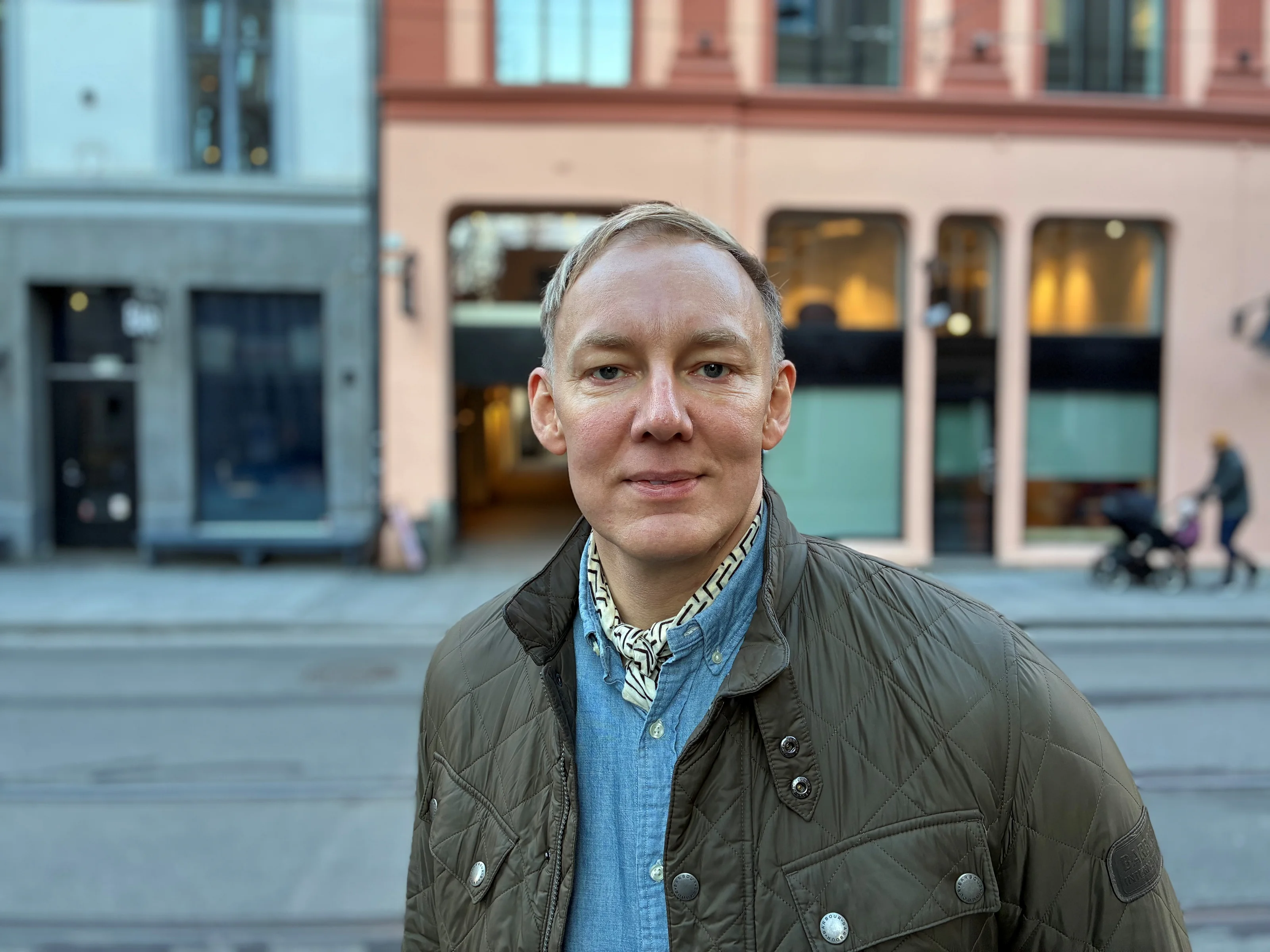 Mann med lyst kortklipt hår ser rett i kamera og smiler. I bakgrunnen er det en rosa bygning og en far som triller en barnevogn.