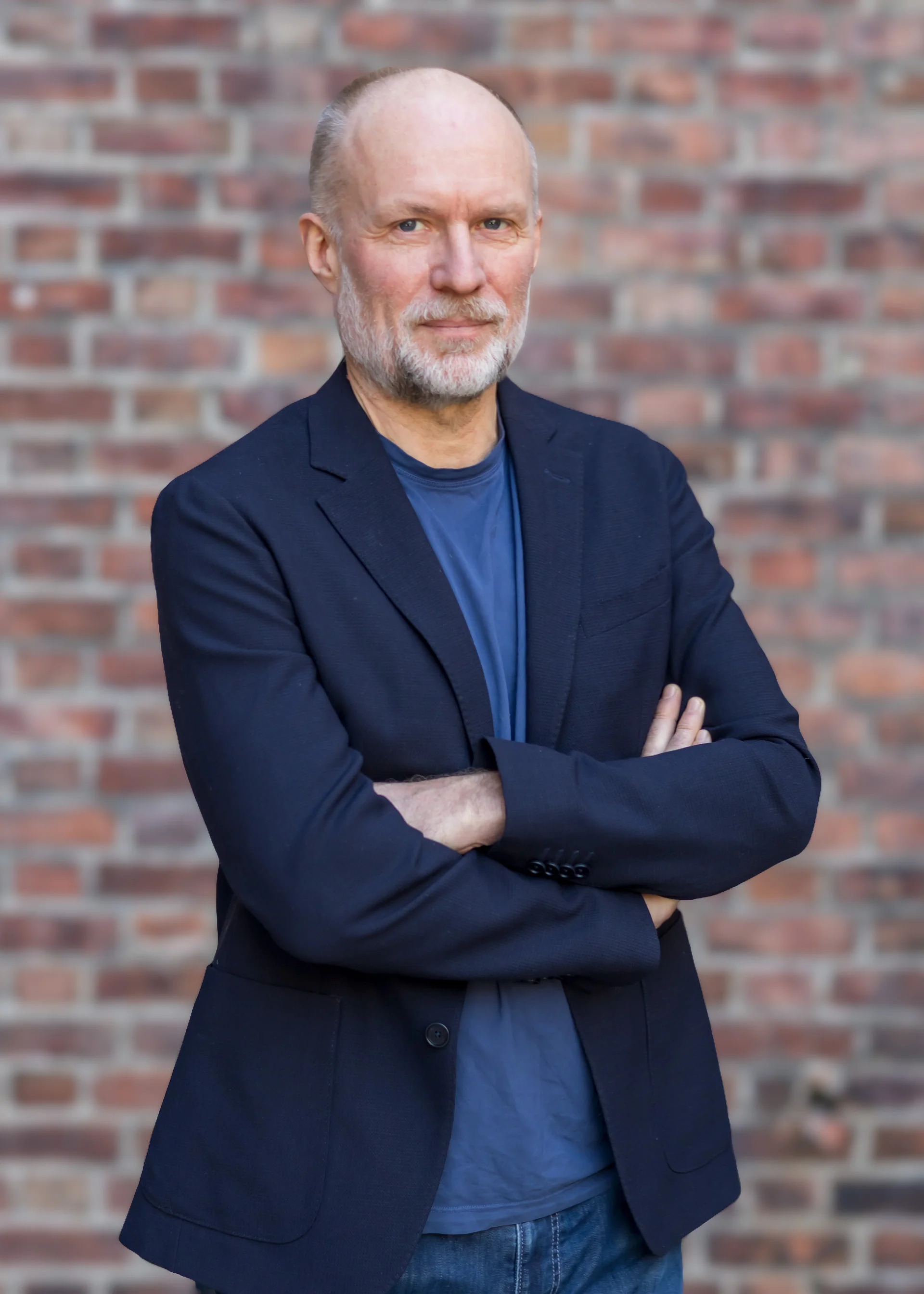 Voksen mann med blå dressjakke og korslagte armer mot en rød teglsteinsmur.