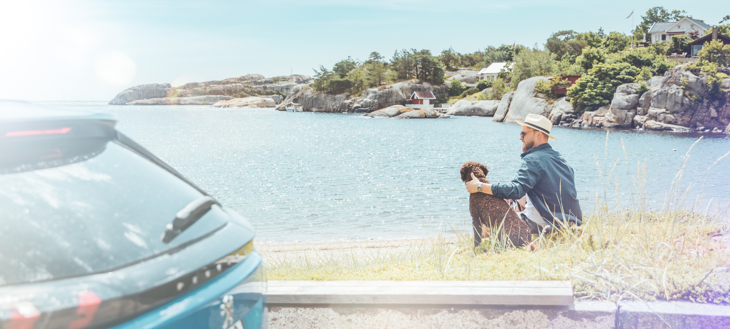 En mann med hund slapper av i strandkanten med bilen parkert i forgrunnen.