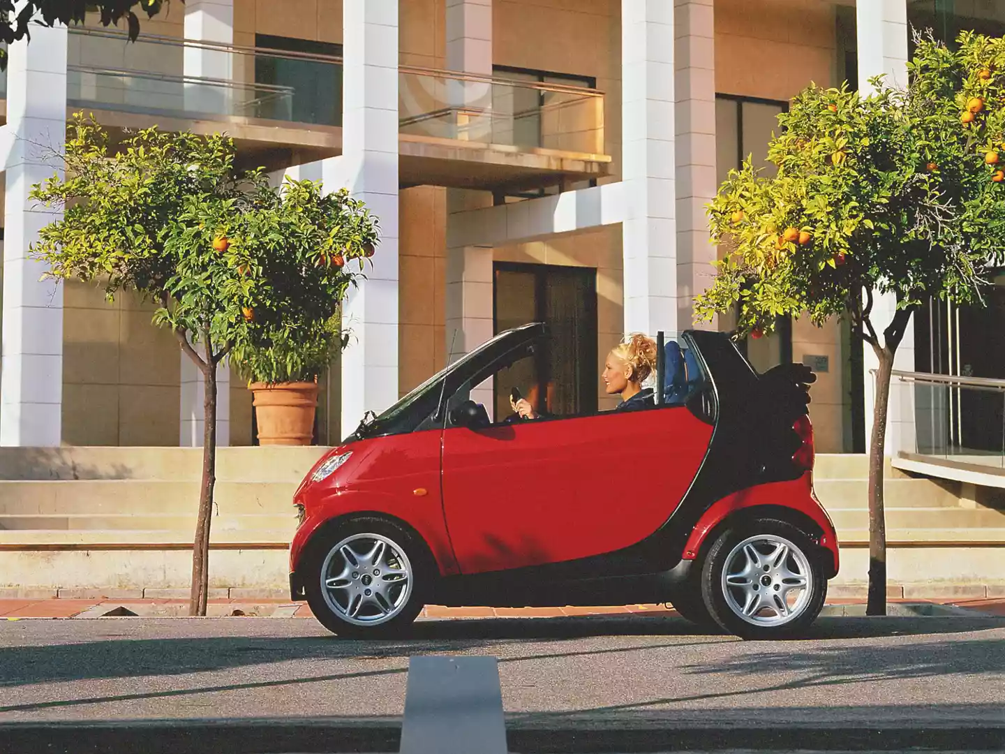 Rød smart fortwo cabrio parkert i bygate mellom appelsintrær