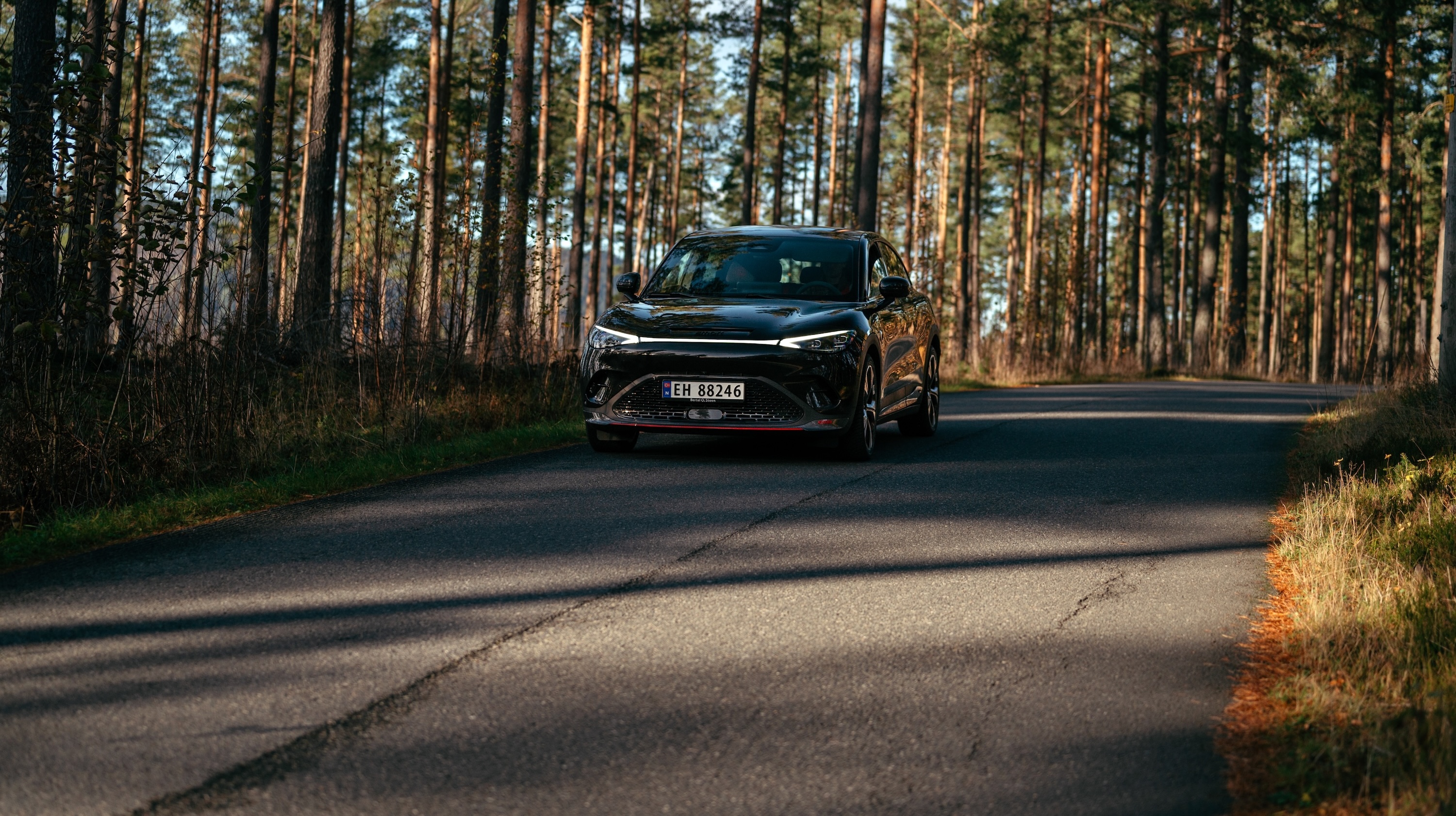 Svart smart #3 BRABUS sett skrått forfra kjører på en svingete asfaltvei gjennom skogen i høstsol