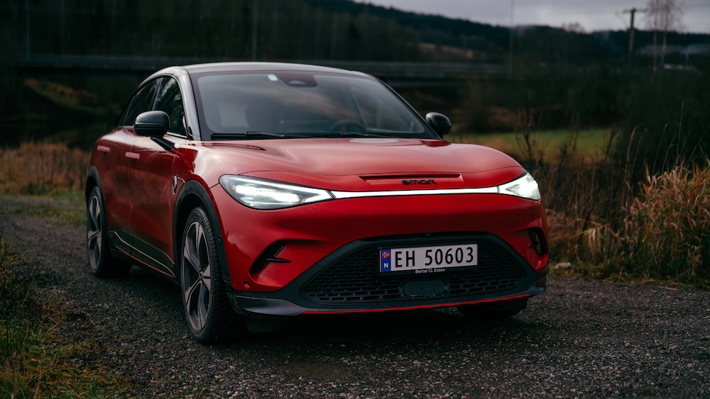 Rød smart #3 BRABUS med svart tak og frontlysene på, sett skrått forfra på en grusvei i landlige omgivelser