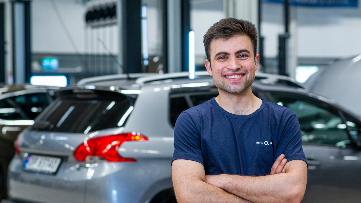 Mekaniker på bilverksted som smiler foran en sølvgrå Peugeot på service i verkstedhallen.