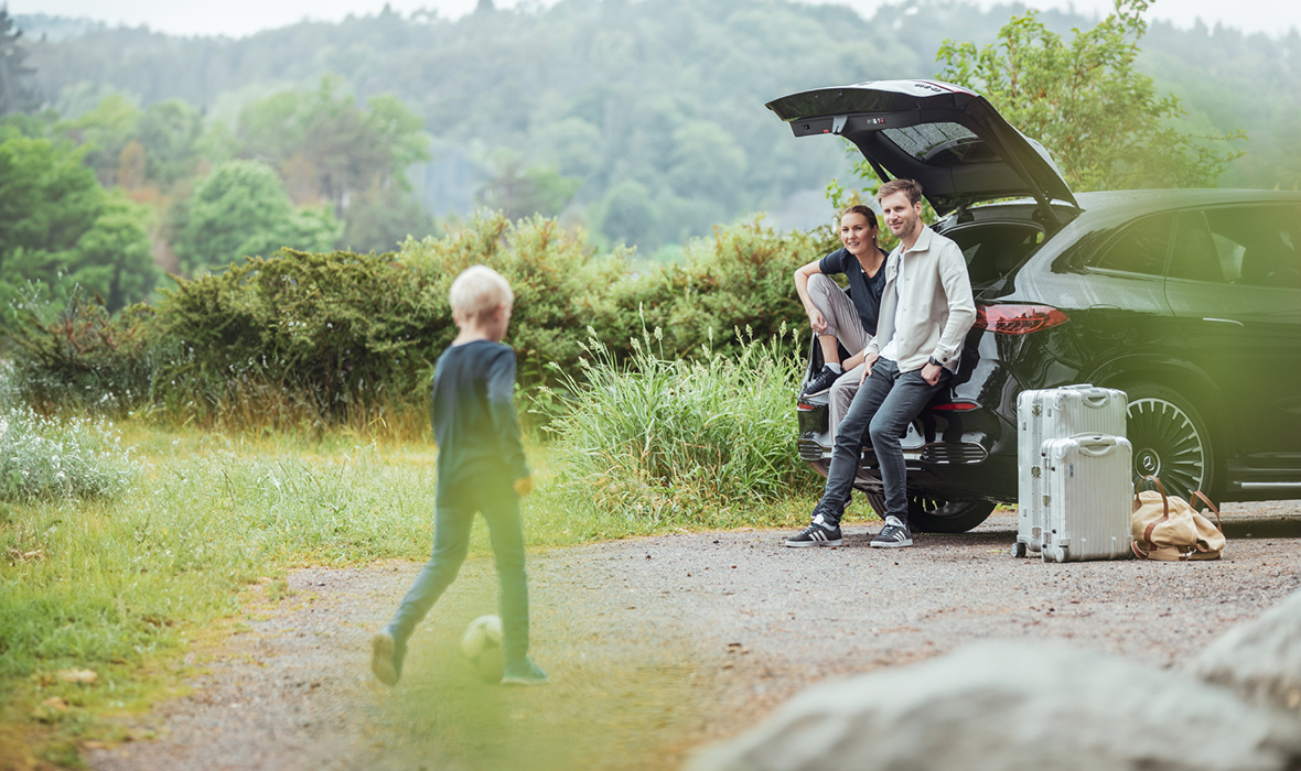 Smilende par med barn i sommerlige omgivelser med Mercedes-Benz EQE SUV i bakgrunnen.