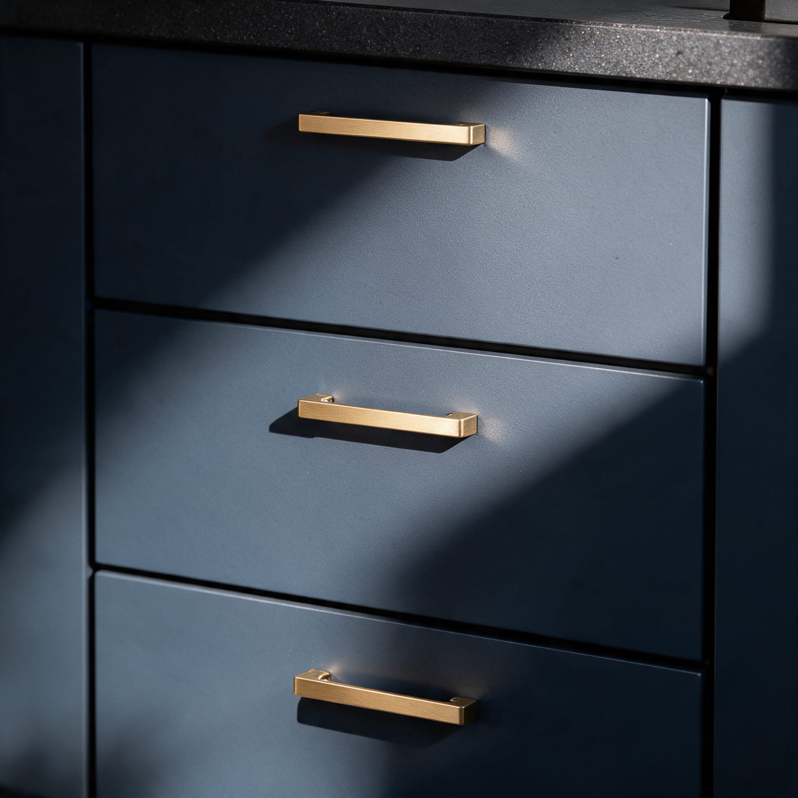 Brushed brass bar handles on deep ink-blue matte drawer fronts below a dark granite worktop.