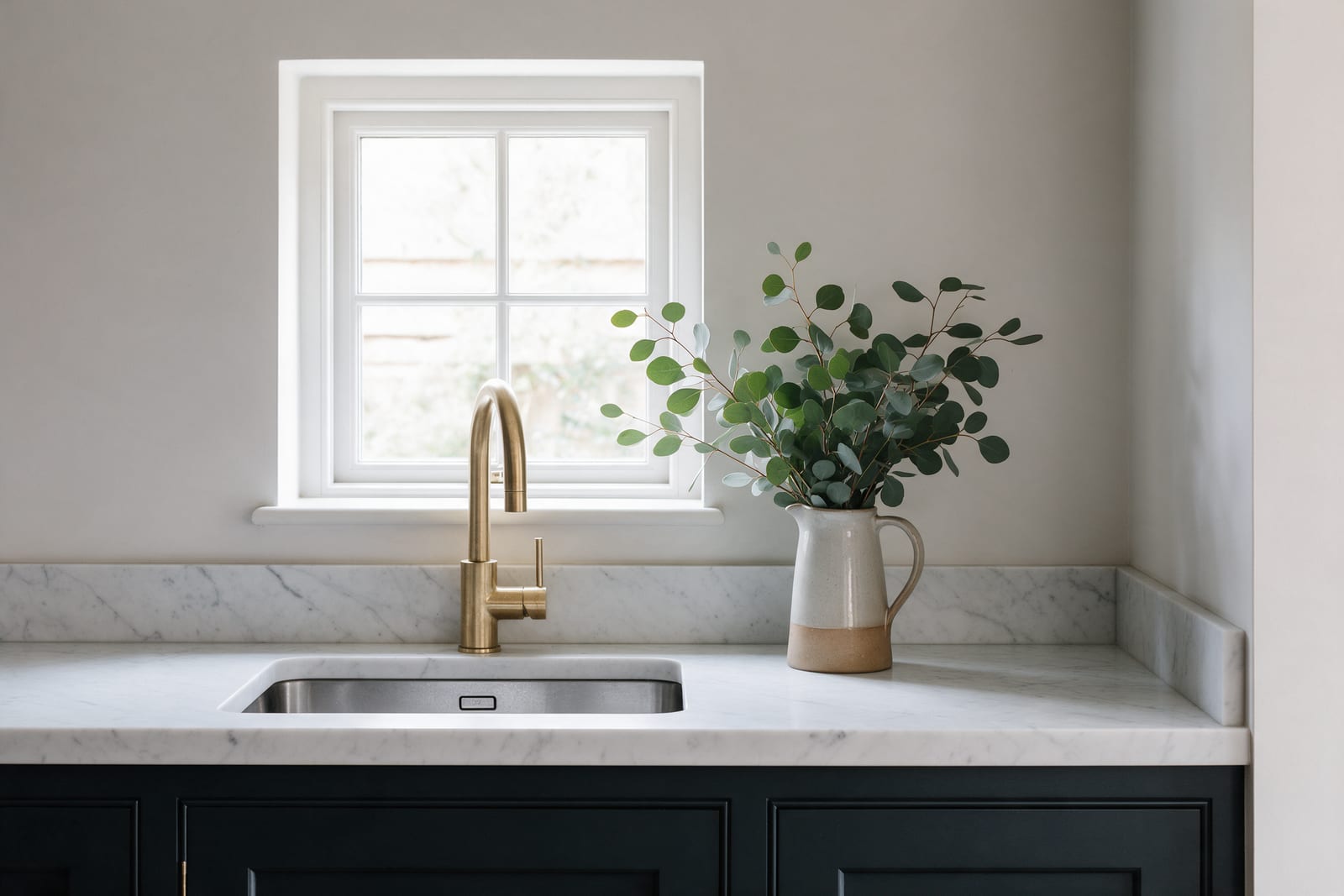 A marble worktop under a small window with a steel sink, brass tap and a jug of eucalyptus.
