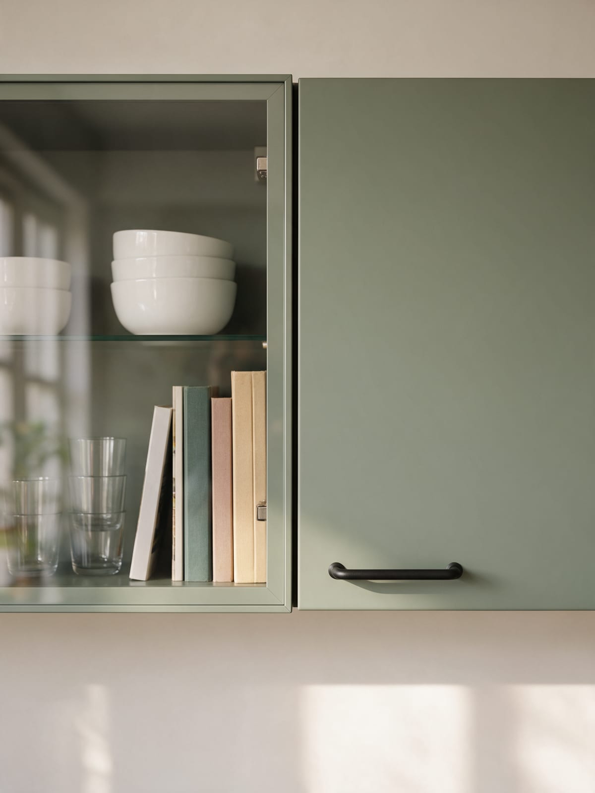 A glazed wall cabinet with white bowls, glasses and cookbooks beside a sage green wall cabinet.