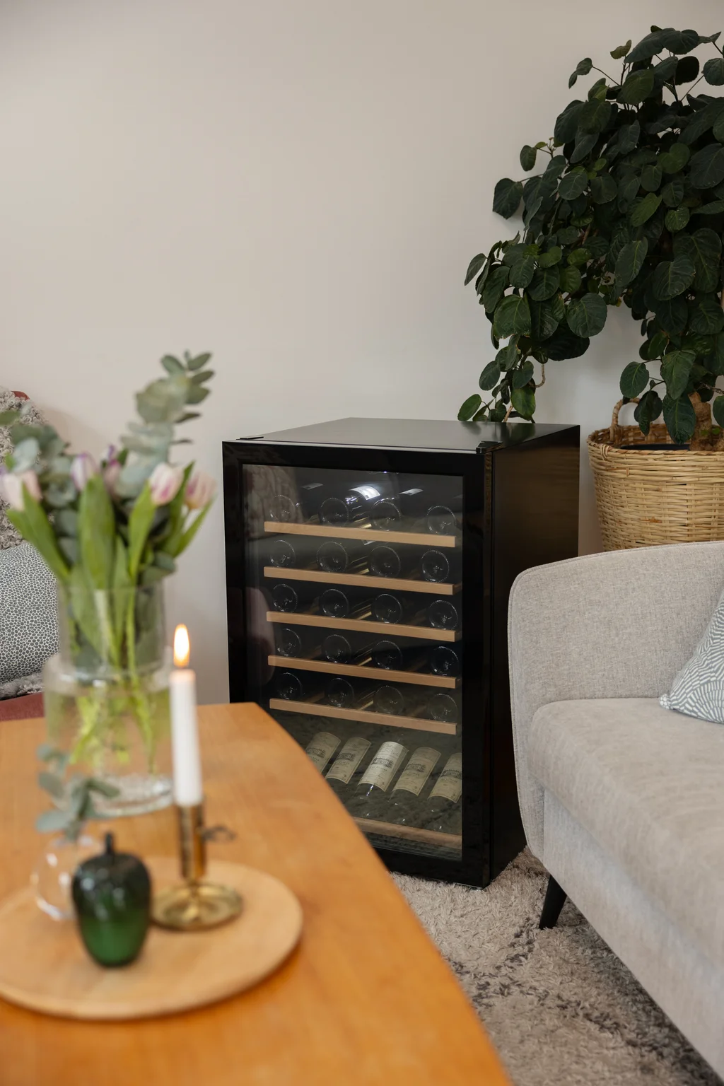 Sort vinkøleskab fra Cavin med glasdør og træhylder i en stue, mellem en beige sofa og et sofabord med tulipaner og et stearinlys