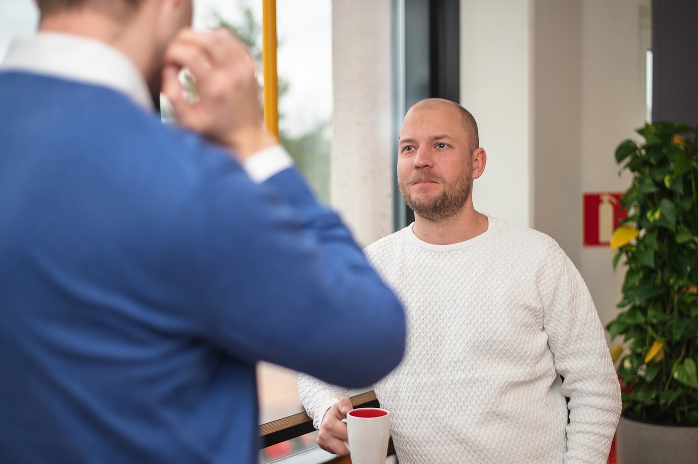 Två kollegor i samtal, där en man i vit tröja håller en kopp. Denna informella interaktion kan bidra till en positiv arbetsmiljö som stöds av Mitsubishi Electrics effektiva klimatlösningar för komfort.