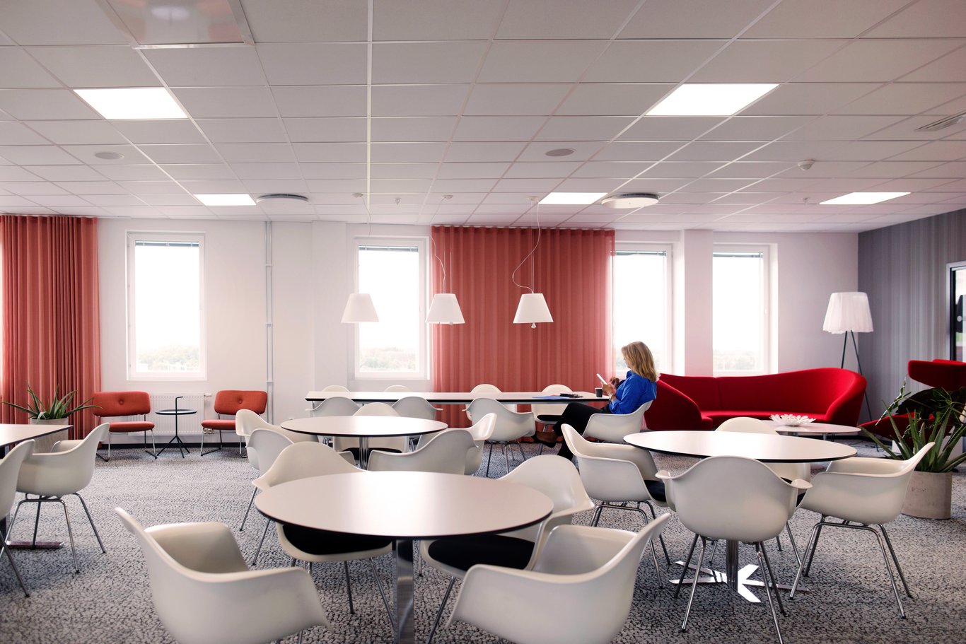 En modern och luftig pausmiljö med vita bord och stolar, samt en röd soffa, där en person sitter och arbetar i ett lugnt kontorsutrymme. Mitsubishi Electric bidrar med energieffektiv ventilation för en behaglig miljö.