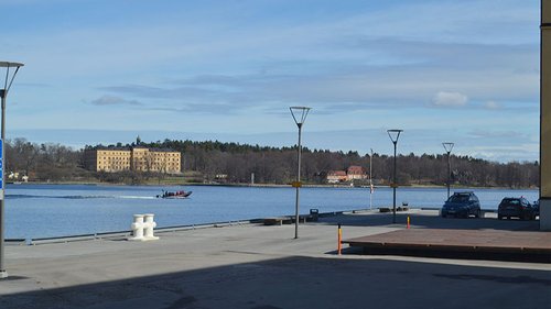 Utsikt över en kaj med vattnet i förgrunden, parkerade bilar, och ett grönskande landskap samt byggnader i bakgrunden. Det är en solig dag med klarblå himmel.