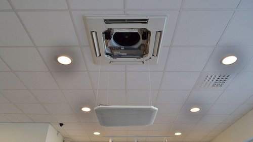 Närbild av en takmonterad luftkonditioneringsenhet med öppnad panel i ett innertak, sannolikt en del av ett ventilations- eller kylsystem från Mitsubishi Electric. Runtom syns taklampor och ett galler för ventilation.