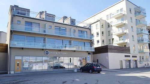 Fasad av byggnad med skylt för "Puls & Träning". Byggnaden har glasfasader och balkonger, och en bil står parkerad framför ingången. Byggnaden ligger i ett område med moderna flerfamiljshus.