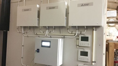 Klimatstyrningssystem från Mitsubishi Electric installerat på väggen, bestående av flera kopplingsboxar och kontrollpaneler. Systemet används för att optimera energiförbrukningen och reglera värme och ventilation effektivt.