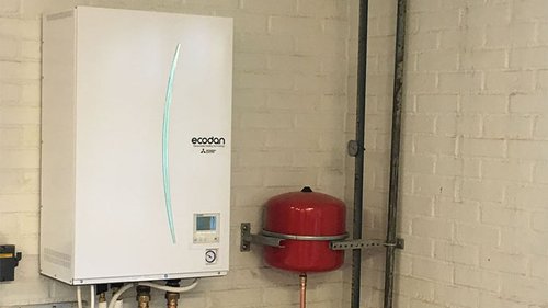 Mitsubishi Electric Ecodan värmepump monterad på en vit tegelvägg, tillsammans med en röd expansionskärl. Systemet är designat för att tillhandahålla effektiv uppvärmning och energibesparing i bostads- och kommersiella fastigheter.