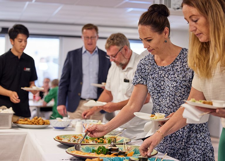 Deltagare från Mitsubishi Electric och andra gäster serverar sig mat från en buffé under ett event i ett modernt kontorsutrymme. Bordet är fyllt med olika maträtter, och människor står i kö för att ta för sig.