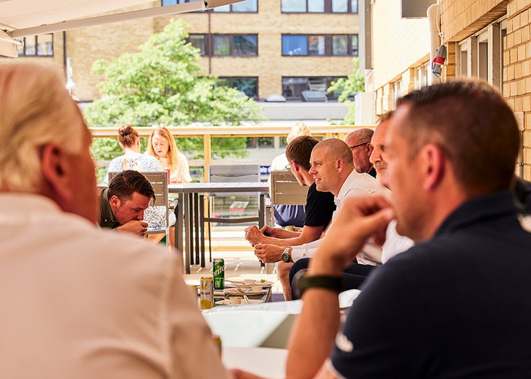 Flera personer sitter och äter på en solig uteplats under ett event, arrangerat av Mitsubishi Electric. De sitter vid olika bord under ett solskydd, med en bakgrund av grönska och närliggande byggnader.