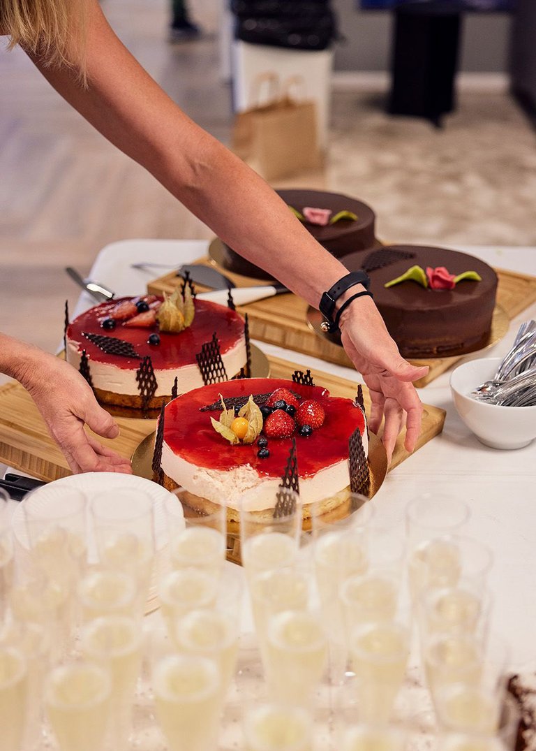 En person placerar en vackert dekorerad tårta med röda bär på ett bord, omgiven av andra desserter. Flera glas med dryck står i förgrunden. Eventet verkar vara en del av ett firande, där Mitsubishi Electric är värd.