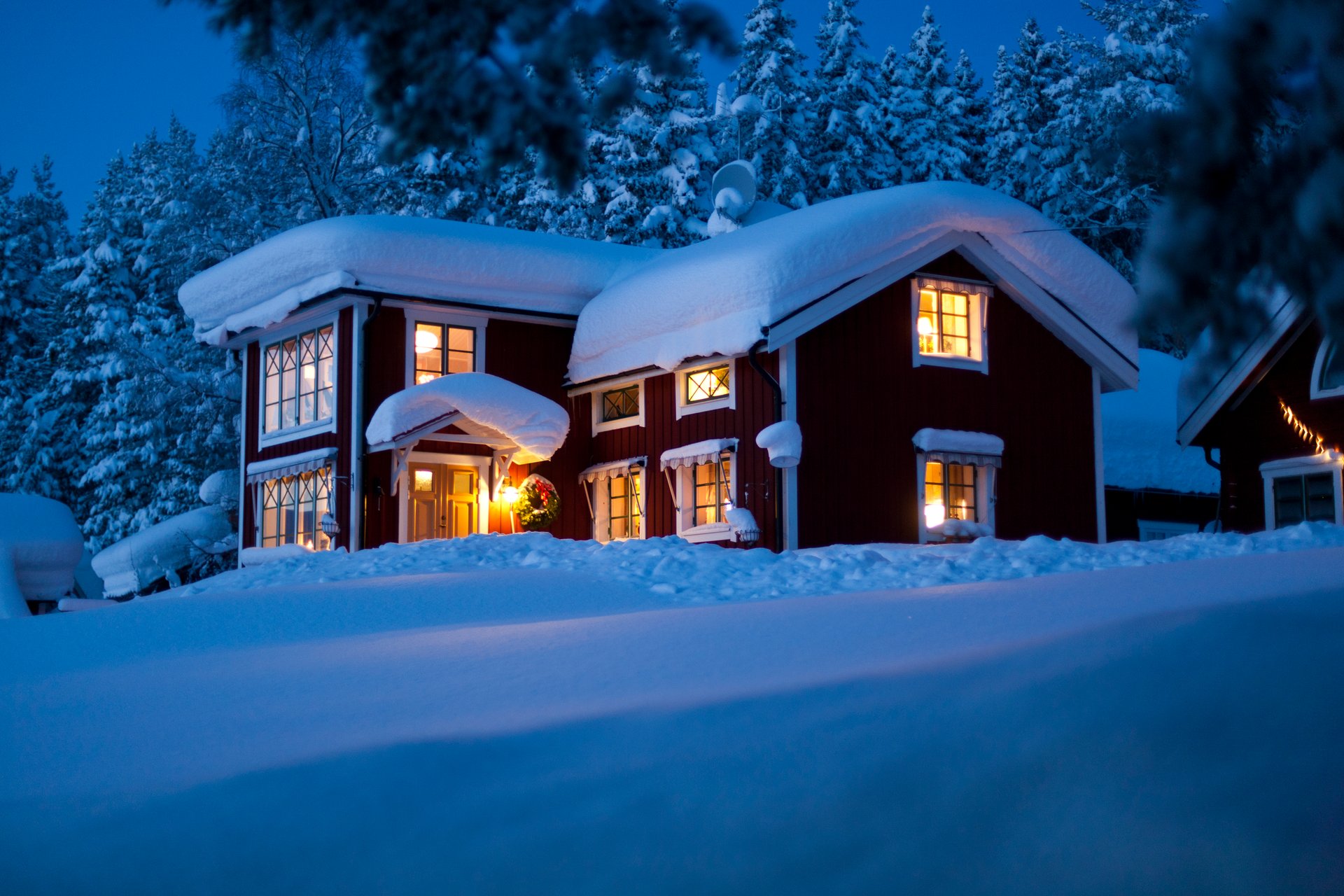 En röd villa täckt av snö på en vinterkväll, upplyst med varma ljus inifrån. Texten bredvid säger "Vi finns från norr till söder. Se filmen om våra installationer runt om i Sveriges avlånga land".