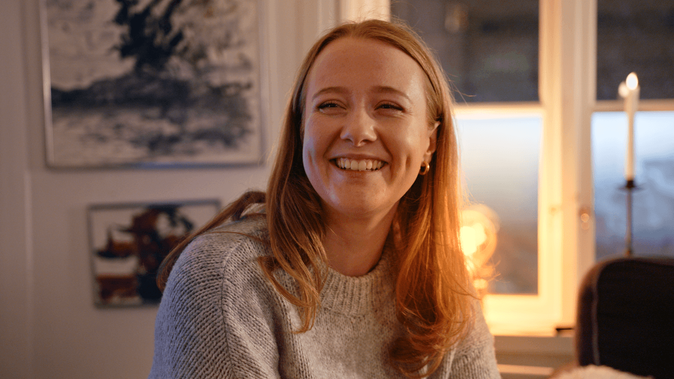 En kvinna klädd i en varm tröja, leende och avslappnad i en mysigt belyst stuga, vilket speglar Mitsubishi Electrics mål att skapa komfort och värme i skandinaviska hem.
