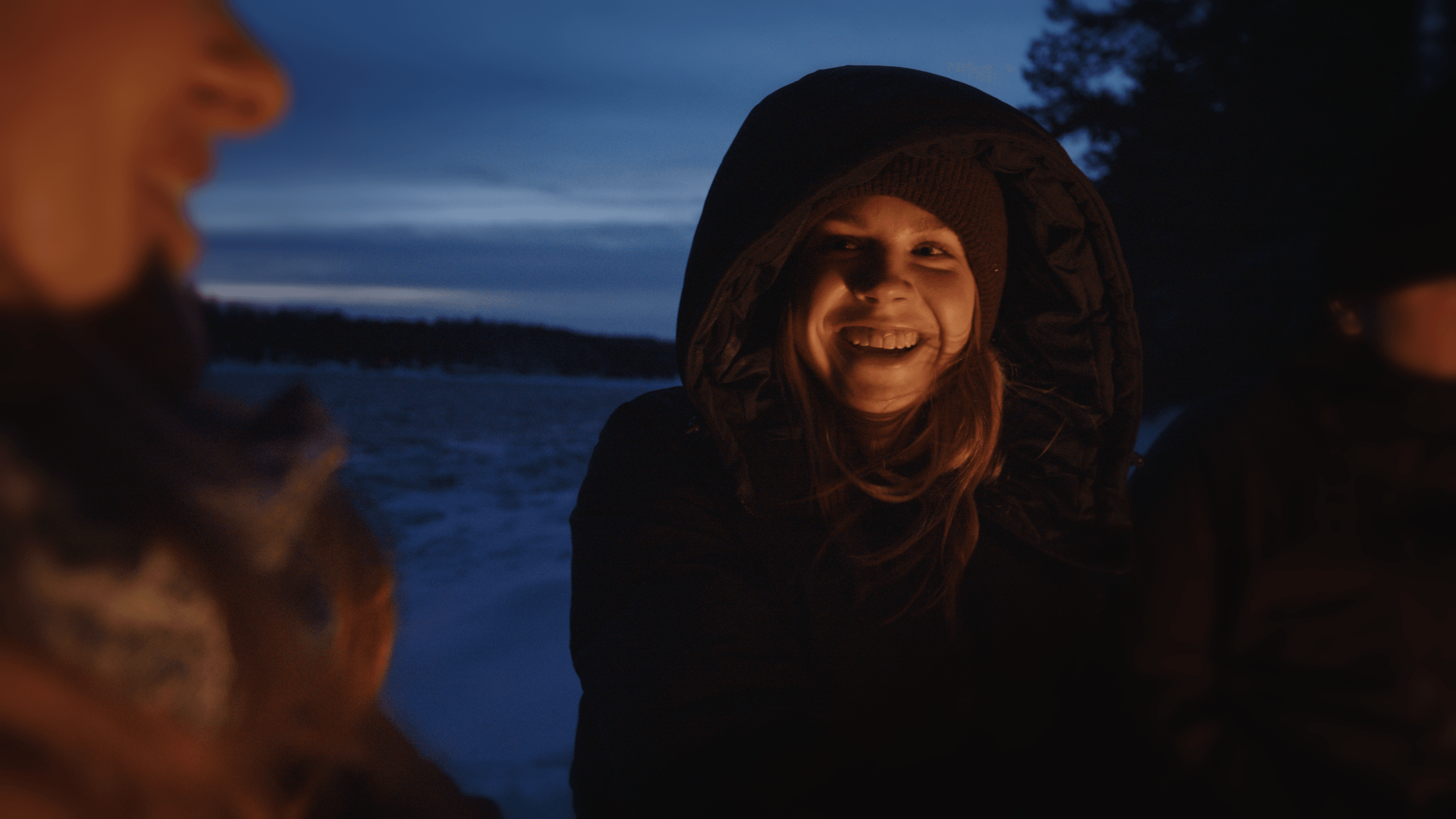 Ett glatt leende från en kvinna i tjock jacka nära en eld, vilket representerar Mitsubishi Electrics uppvärmningslösningar som skapar komfort även i de kyligaste vinterdagarna.