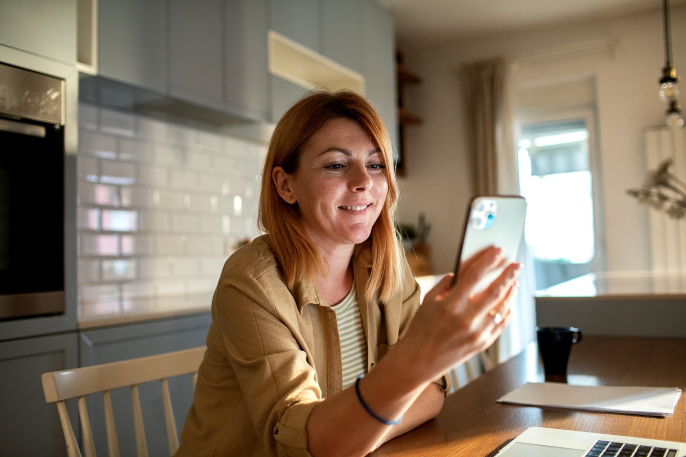 Kvinna använder sin mobiltelefon vid köksbordet hemma.