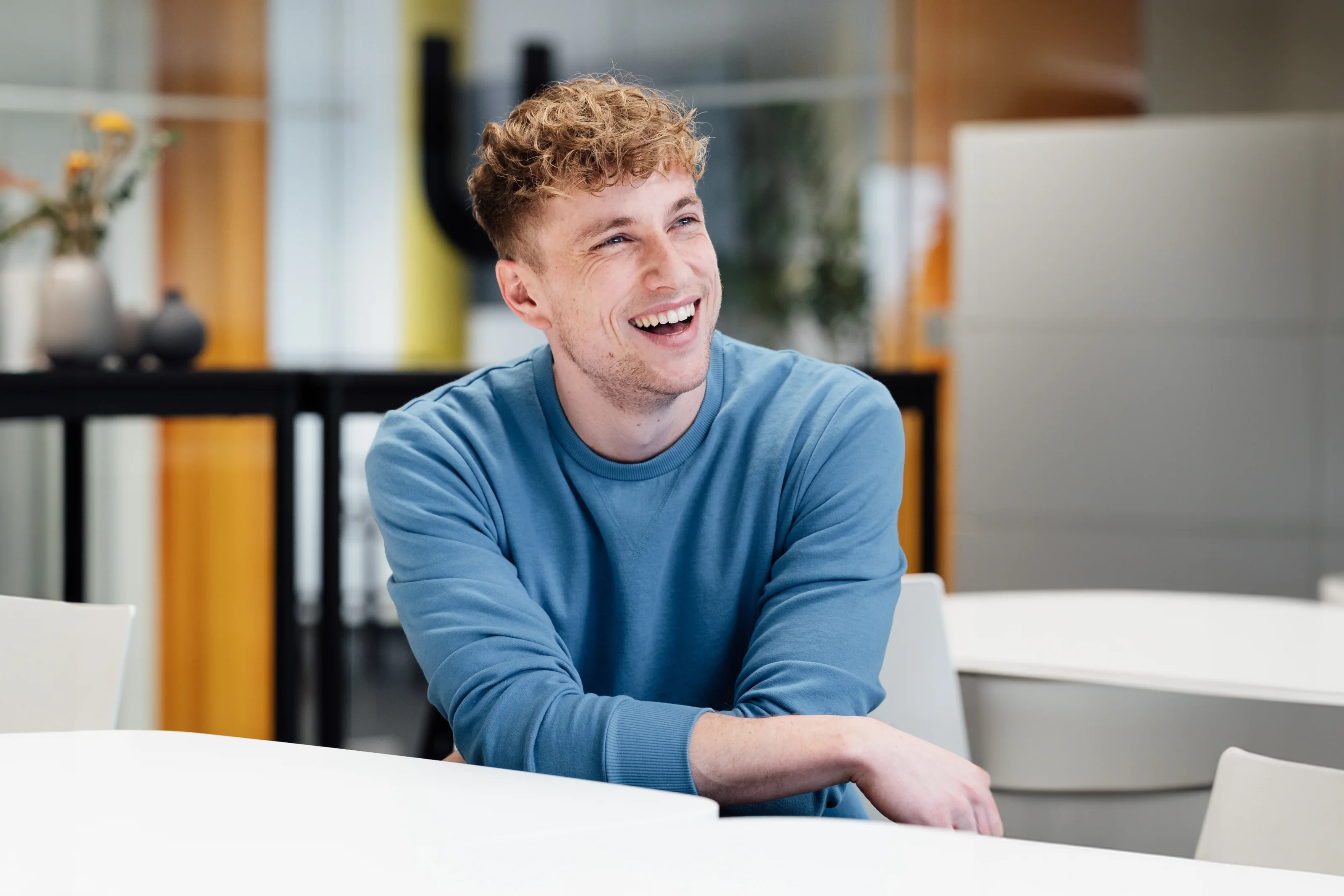 Smiling Nuki team member in a blue sweater sits at a table in the office during a relaxed moment.
