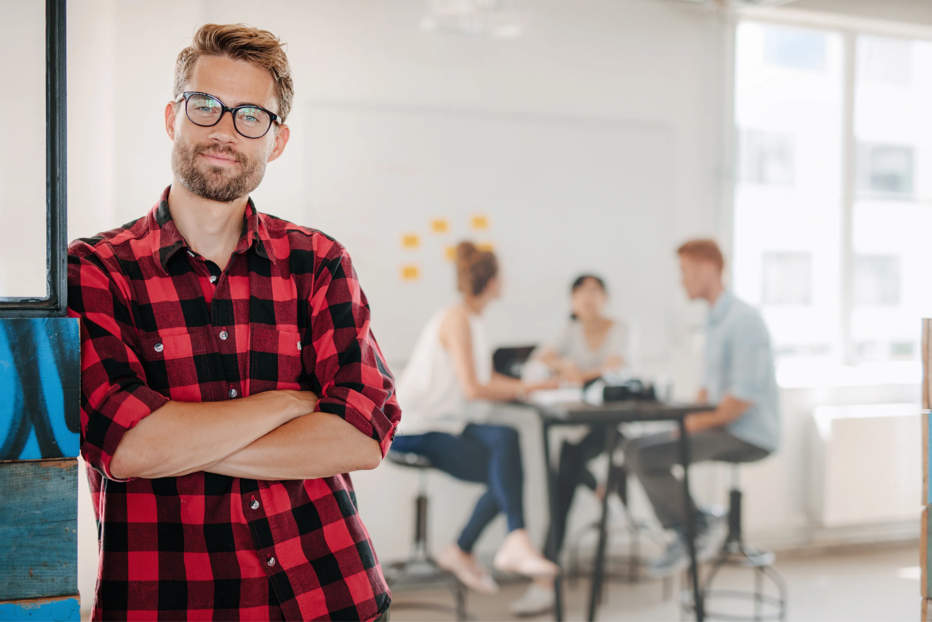 Man in a red plaid shirt leaning against a wall, with a team collaborating in the background.