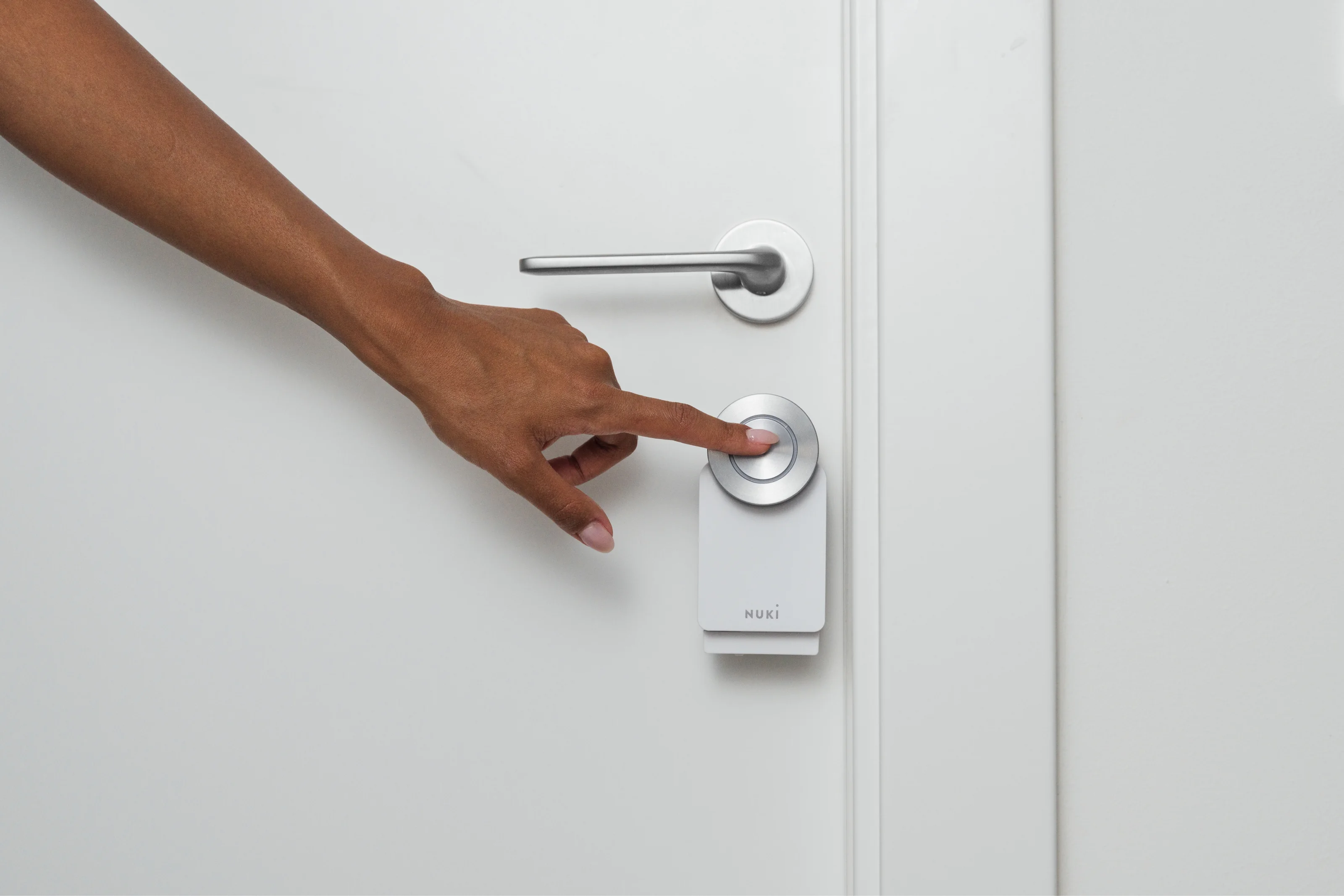 A hand presses the button on the Nuki Smart Lock to unlock a white door effortlessly.