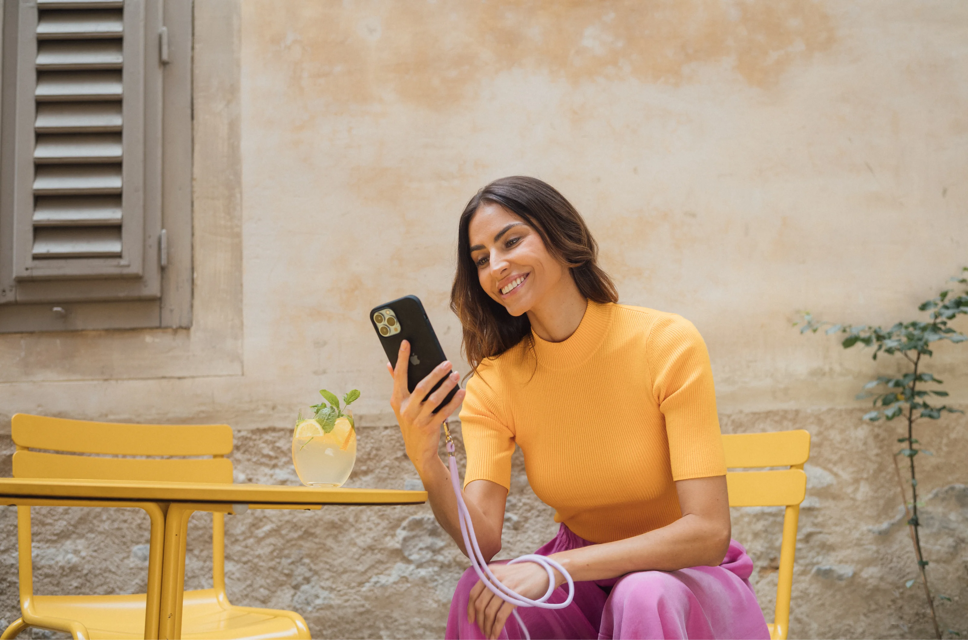 Woman smiles while sitting at a yellow table outdoors, looking at her smartphone in a relaxed setting.