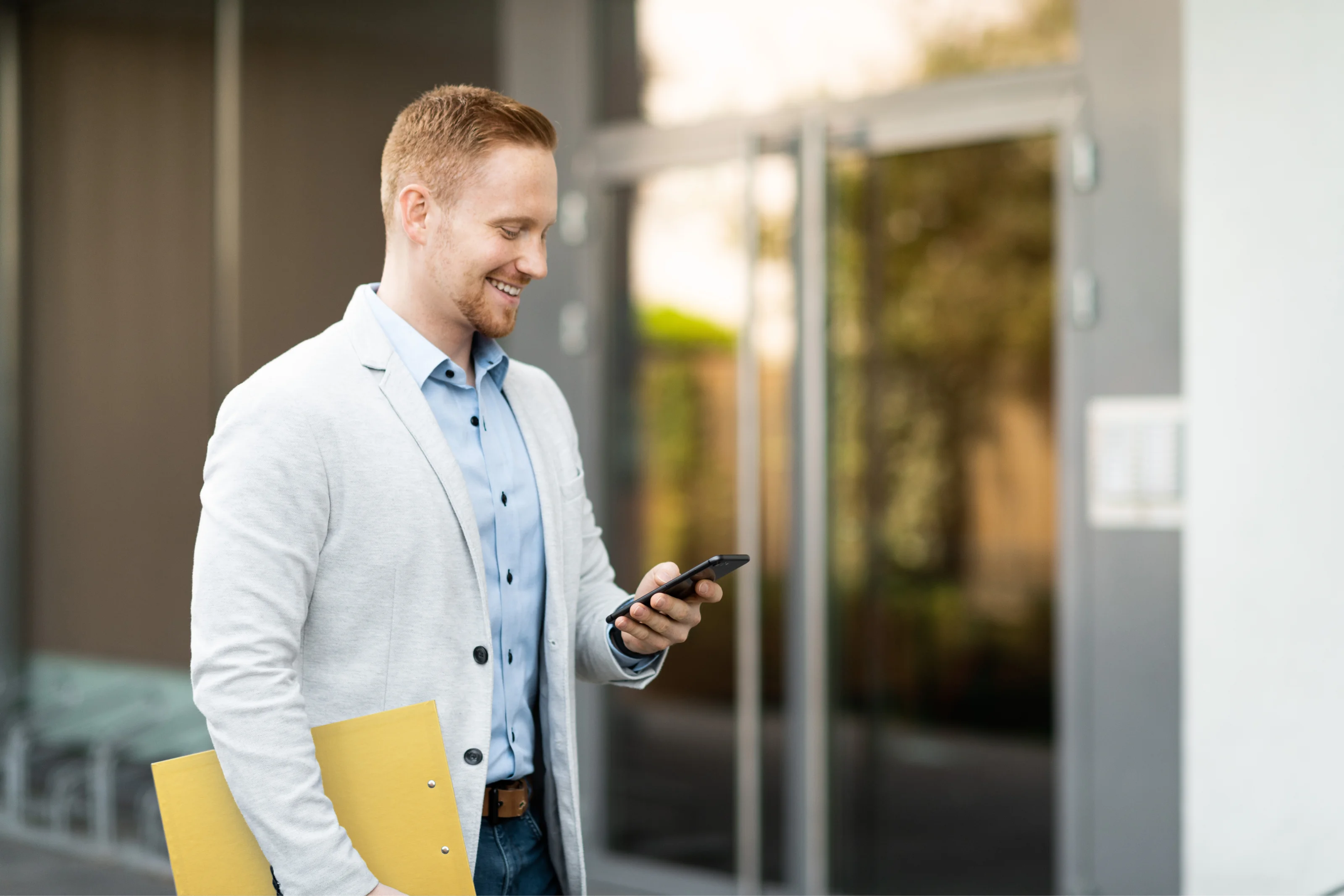 Smiling man in business attire standing in front of a building entrance, looking at his smartphone.