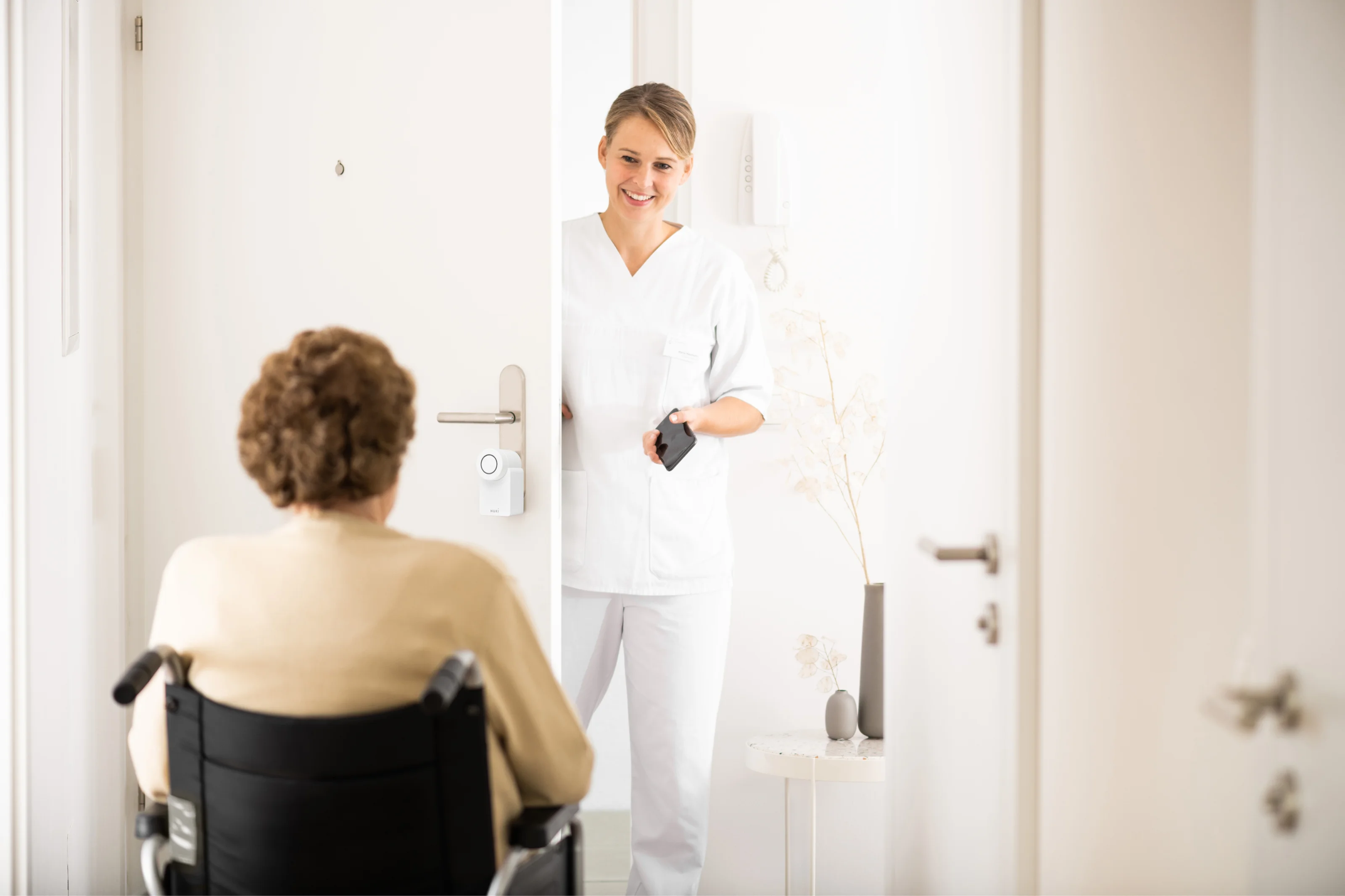 Caregiver opens door with smartphone via Smart Lock while a person in a wheelchair waits inside.