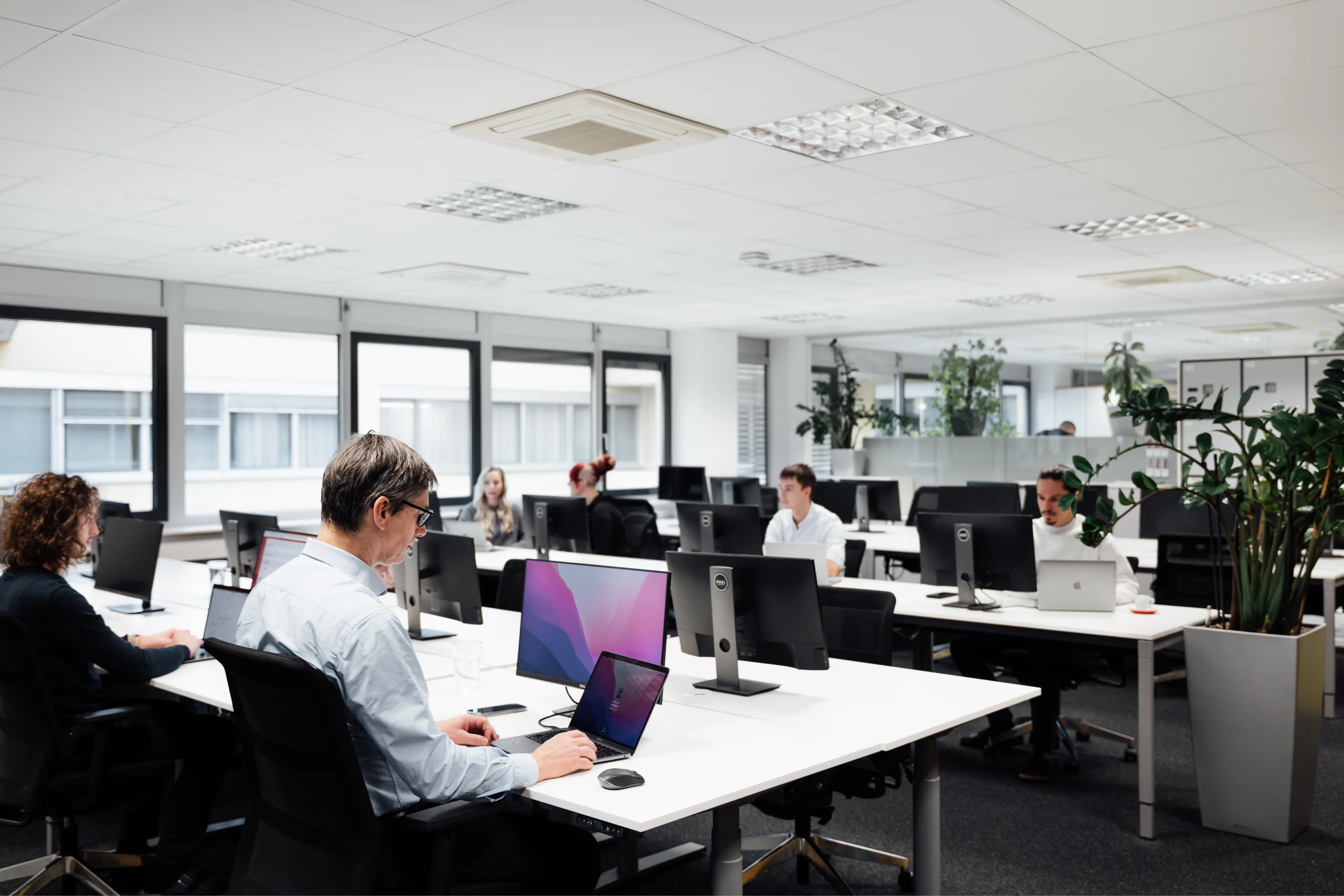 Open office space with people working at desks on computers and laptops in a bright, modern environment.