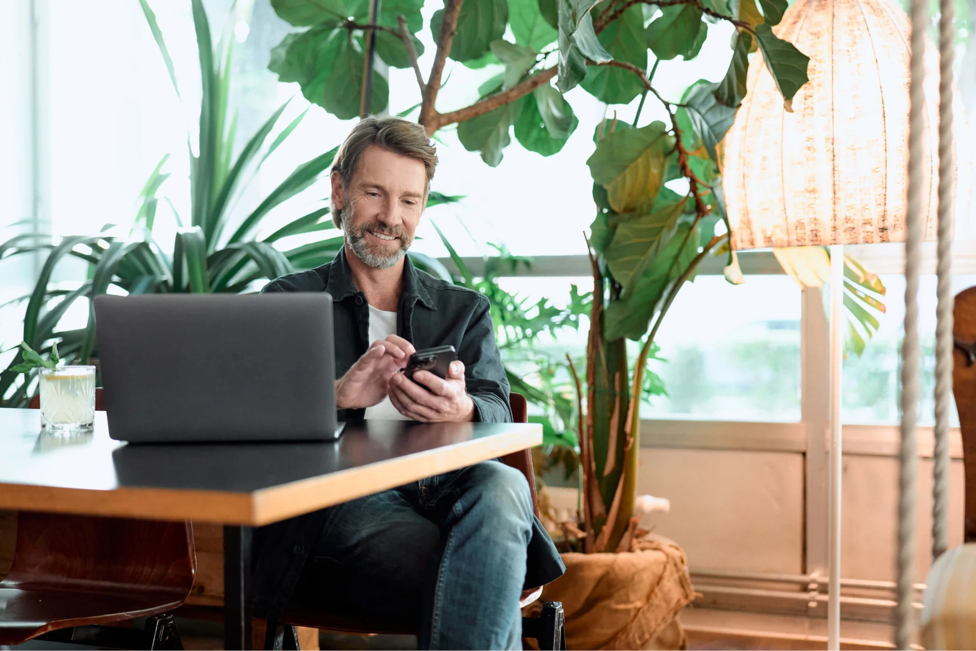 Smiling man using smartphone next to laptop in a cozy, plant-filled workspace