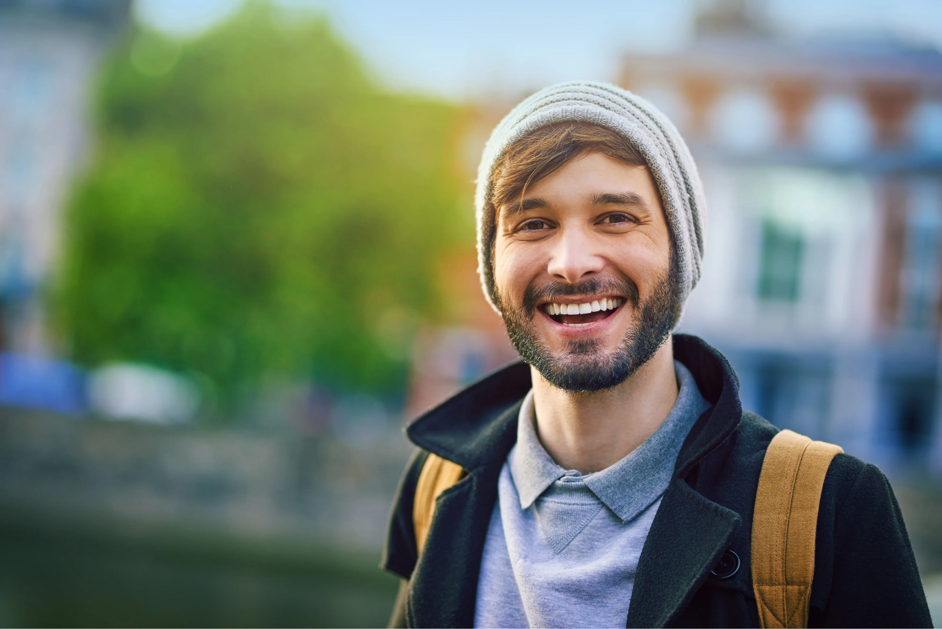 A man with a hat on stands in a city and smiles