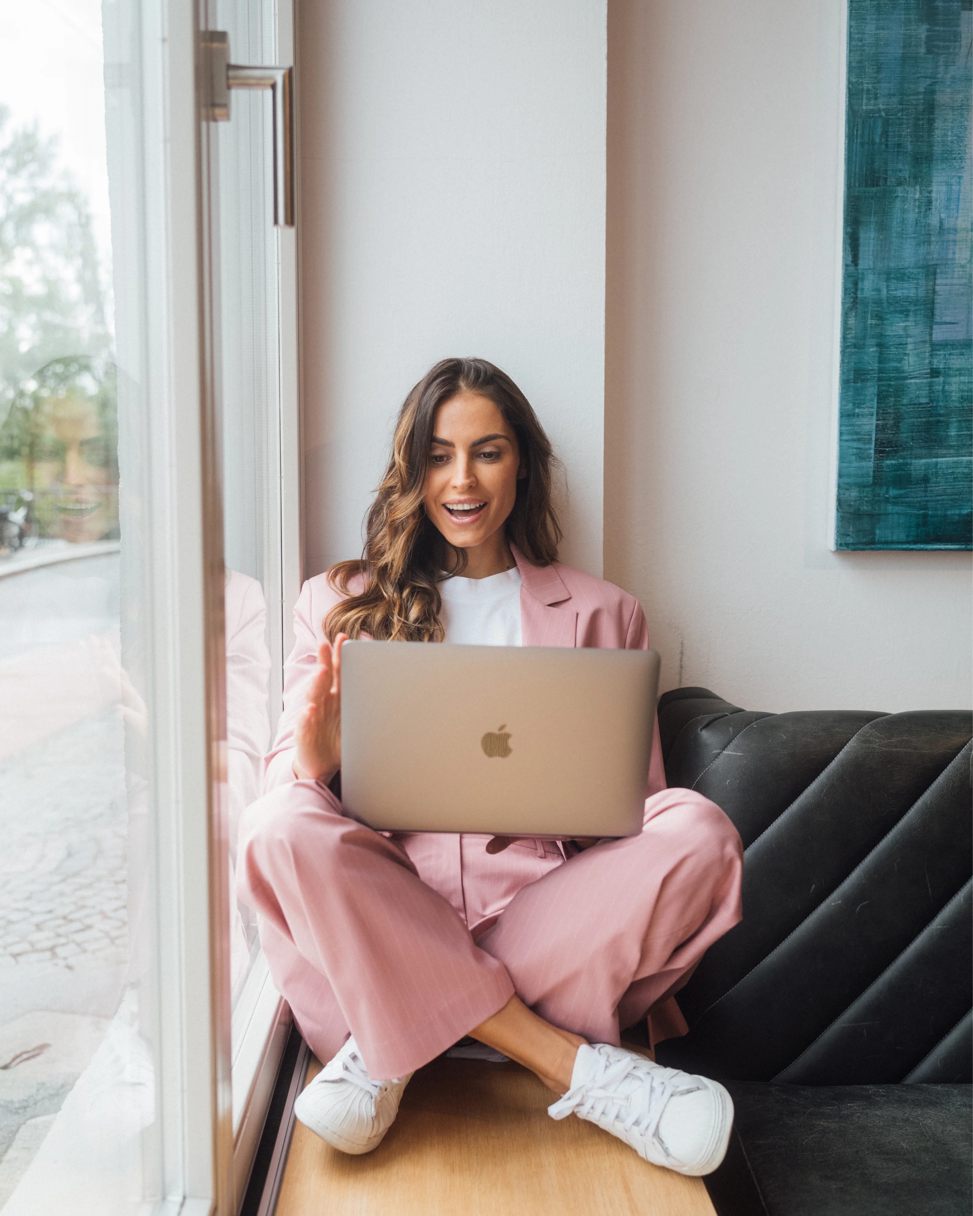 Woman in a pink suit sits on a windowsill with her laptop on her lap.