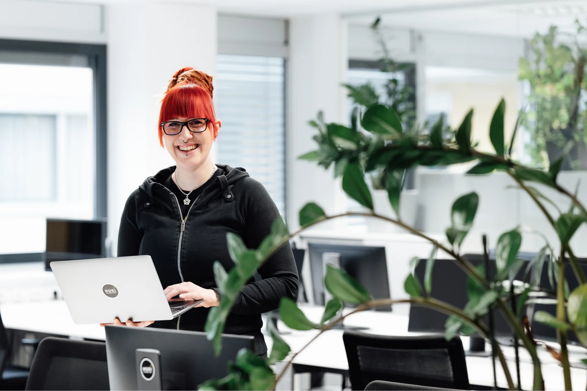 Smiling Nuki team member stands in a bright office holding a laptop with a Nuki sticker, surrounded by plants.