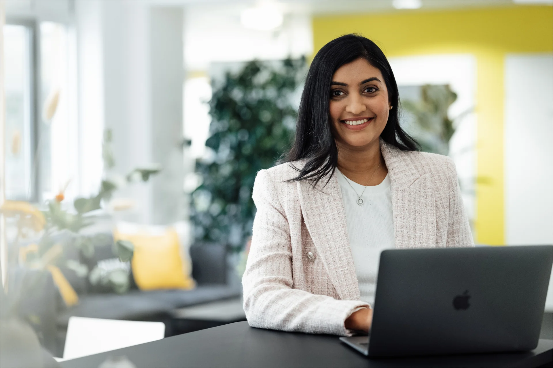 Smiling Nuki team member in a bright office space, wearing a blazer and working on a dark laptop.
