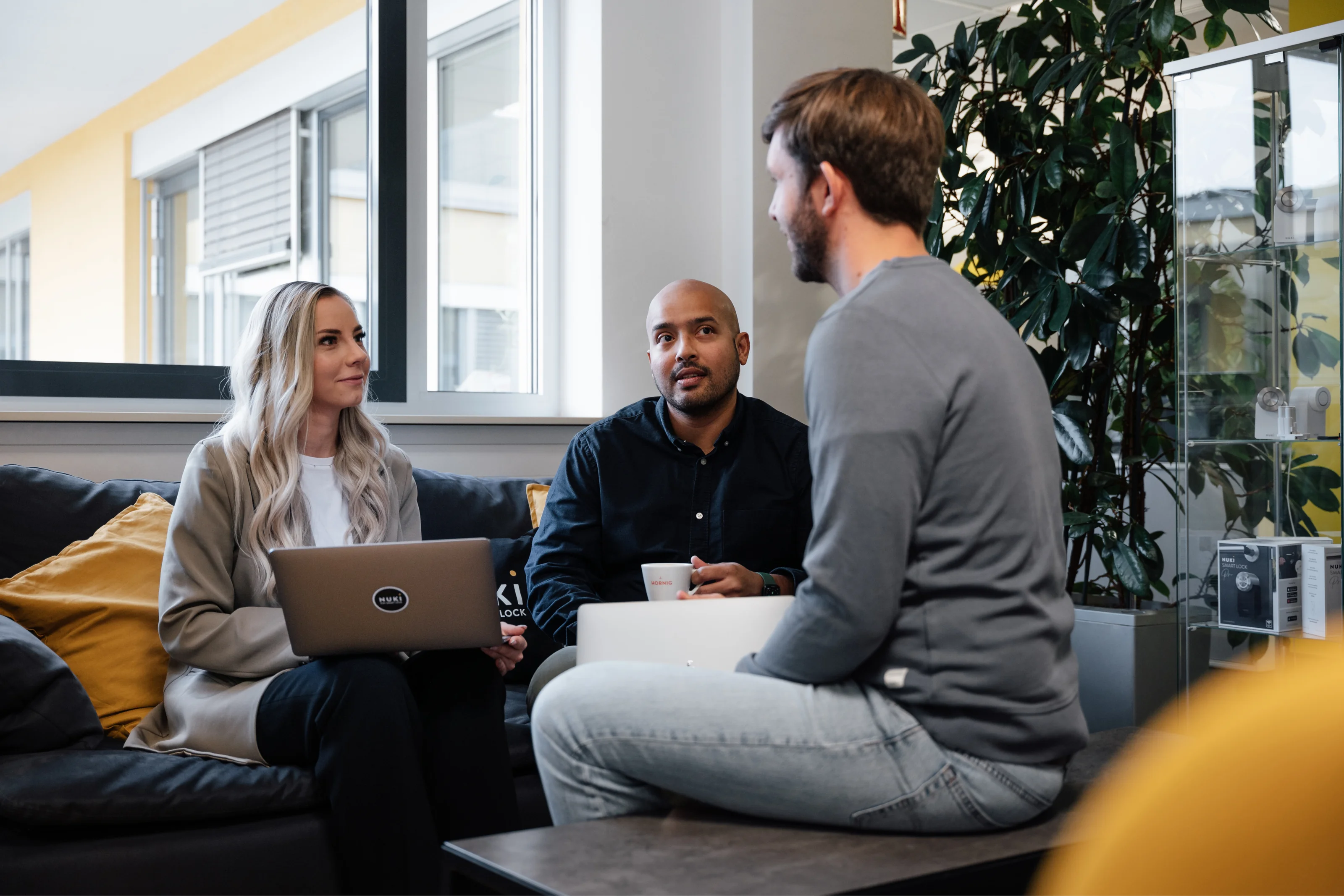Three Nuki team members sit on a couch in a casual office meeting, with laptops and coffee in a bright room.