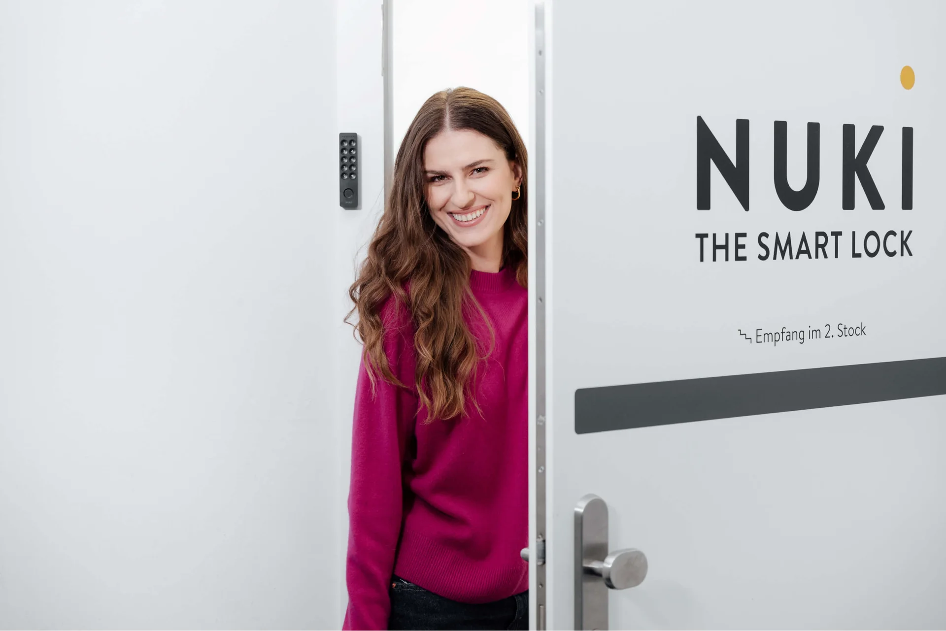 Smiling woman opens the Nuki office door with visible keypad and company logo in a bright entrance area.
