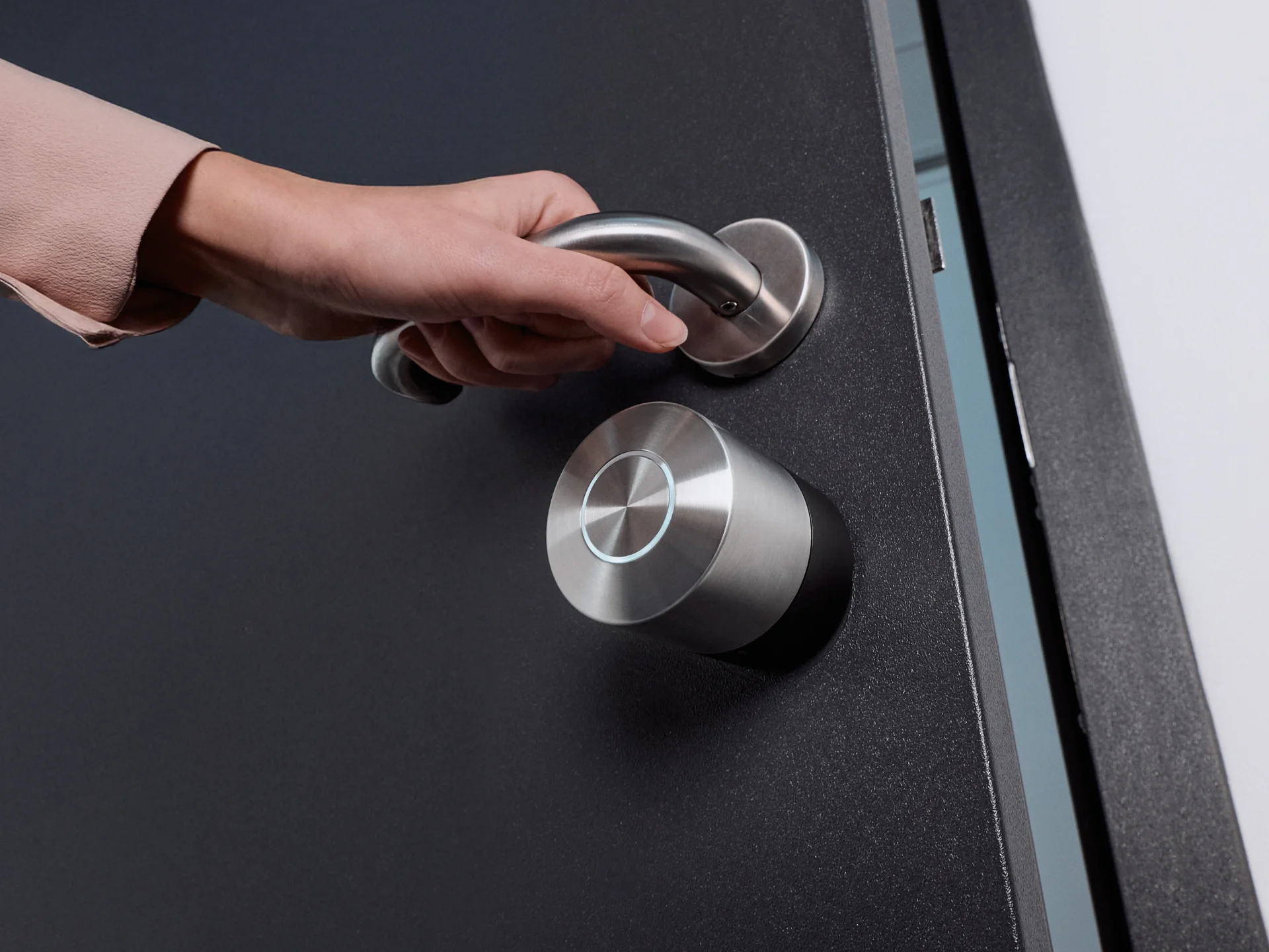 A hand opens a black door with silver hardware with a NUKI Smart Lock Pro mounted on it.