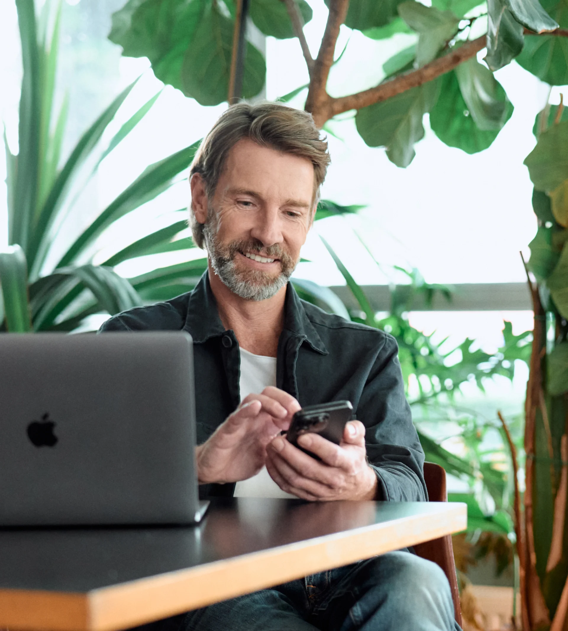 Smiling, middle-aged man with a beard using a smartphone next to a MacBook and surrounded by green plants.