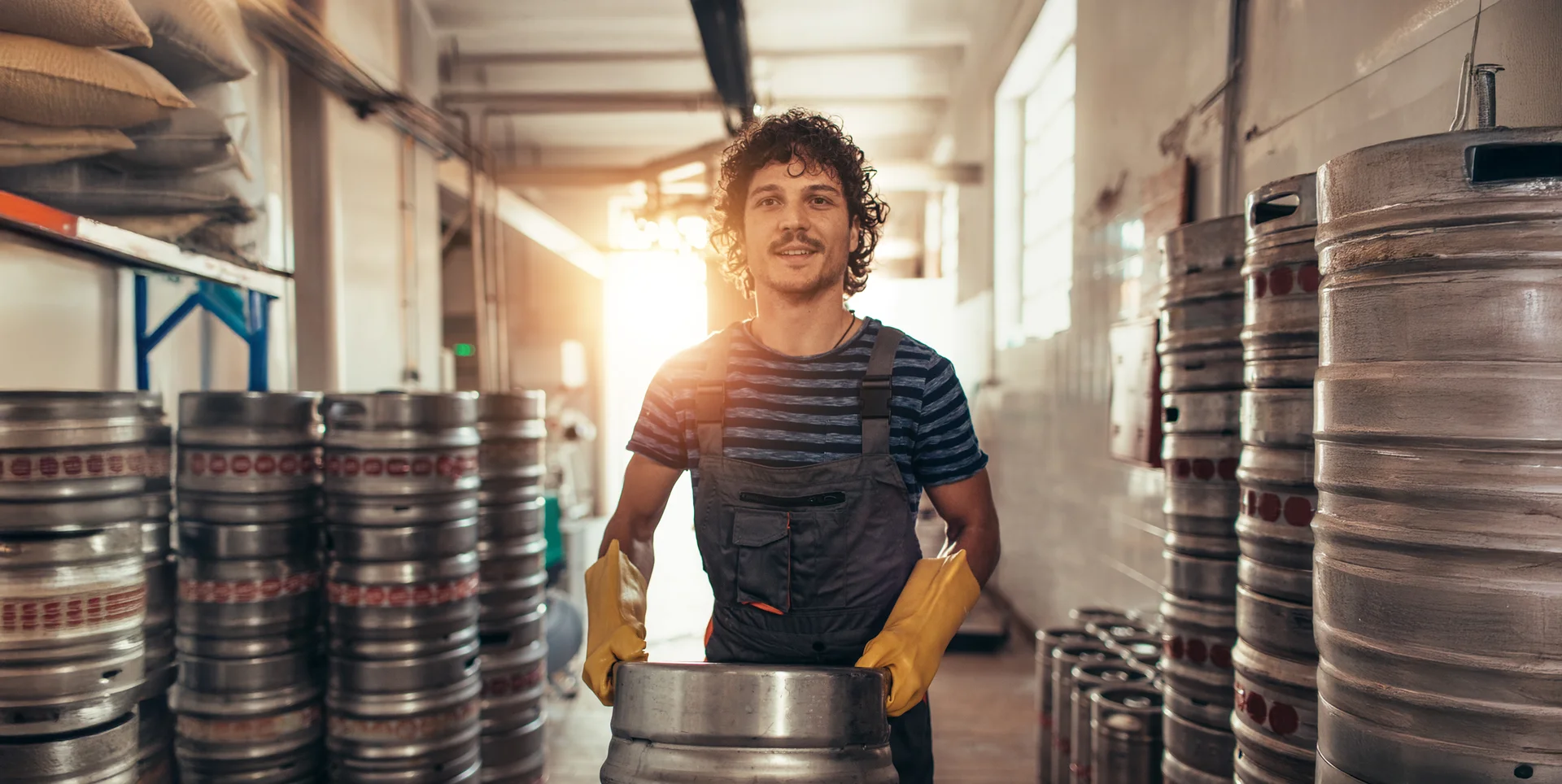 Smiling brewery worker with yellow gloves moving a beer keg in a sunny warehouse, surrounded by stacks of kegs.