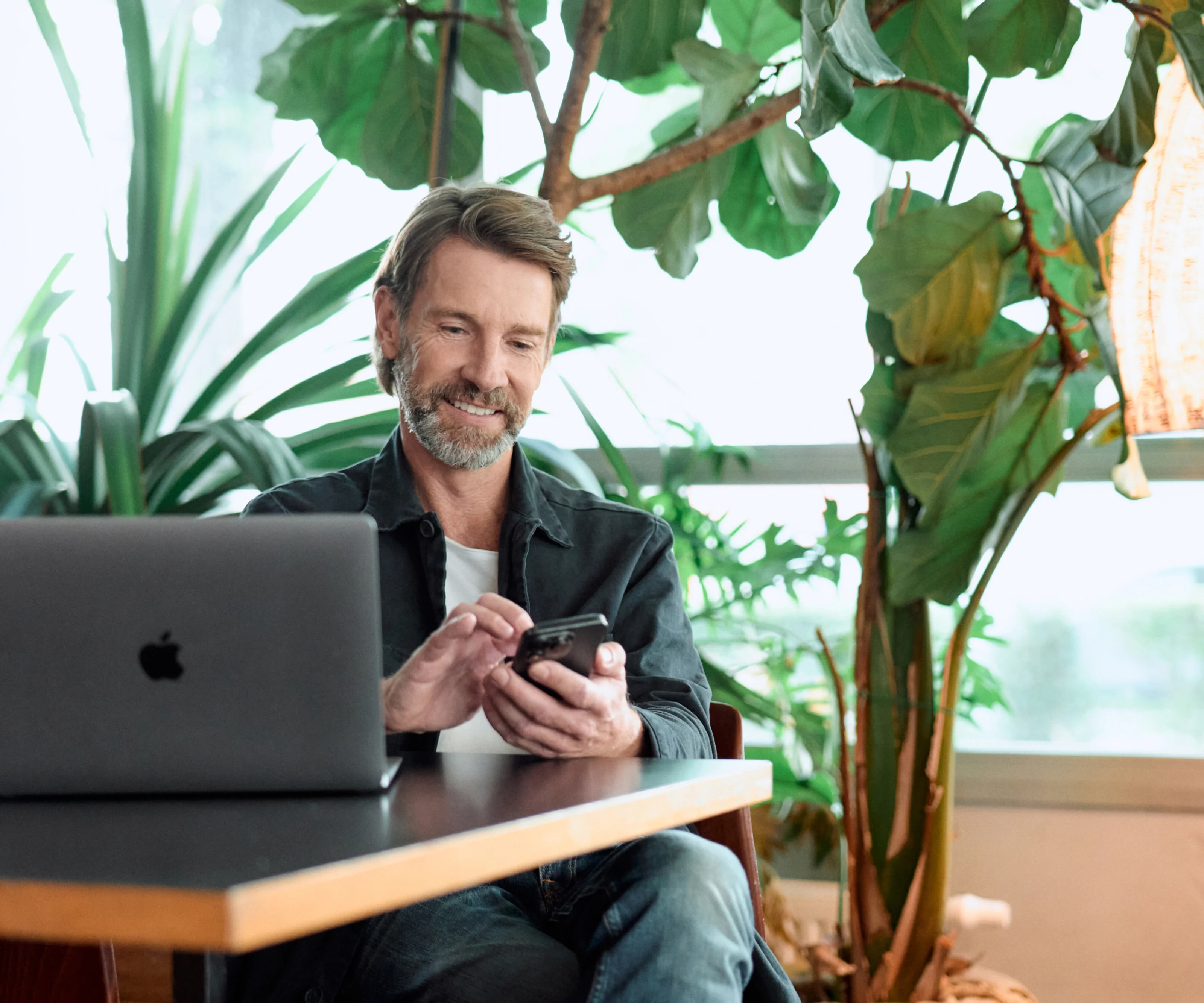 Smiling man working on a laptop and using a smartphone in a bright, indoor setting with large green plants.