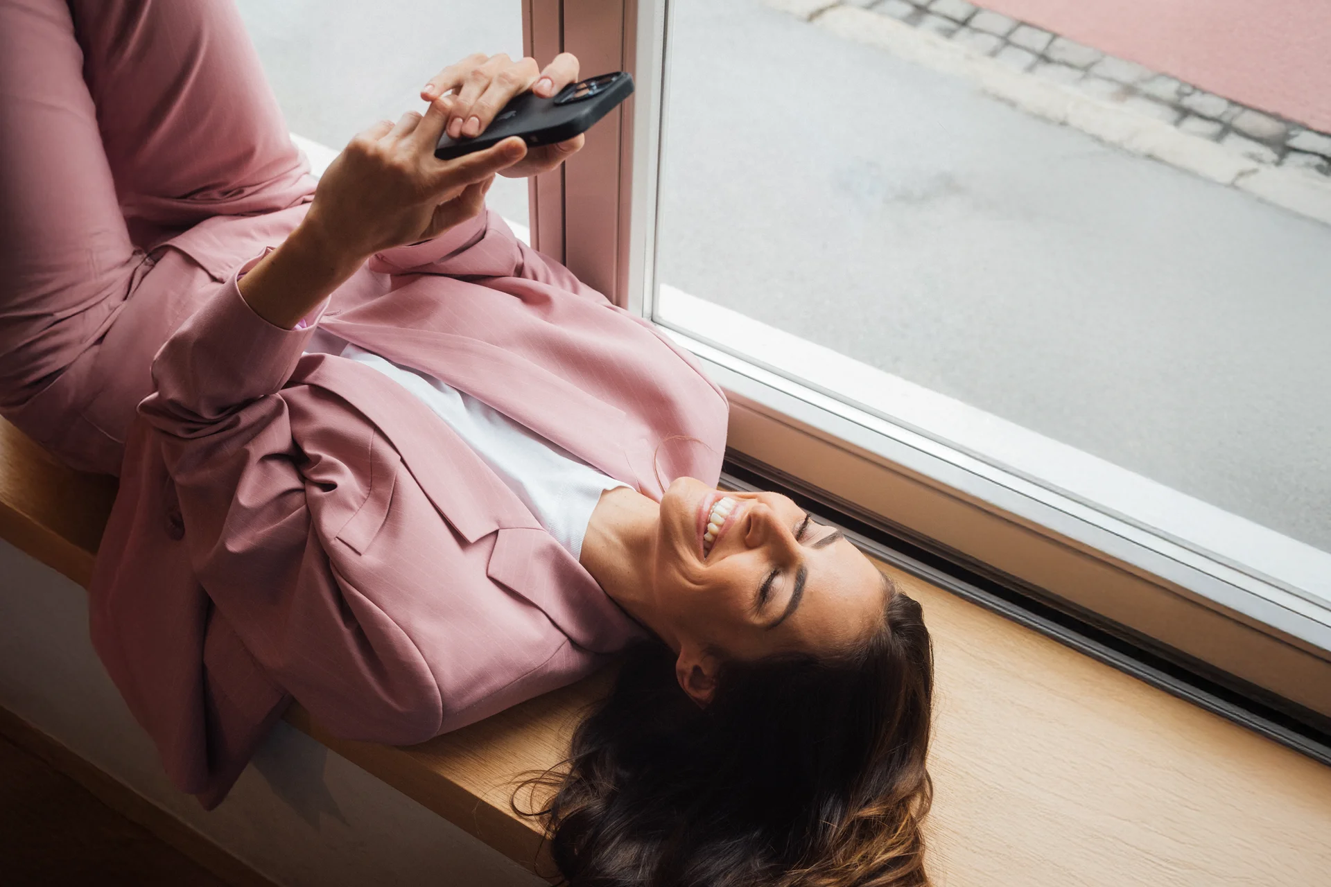A woman lying on a windowsill in a pink suit, looking at her phone.