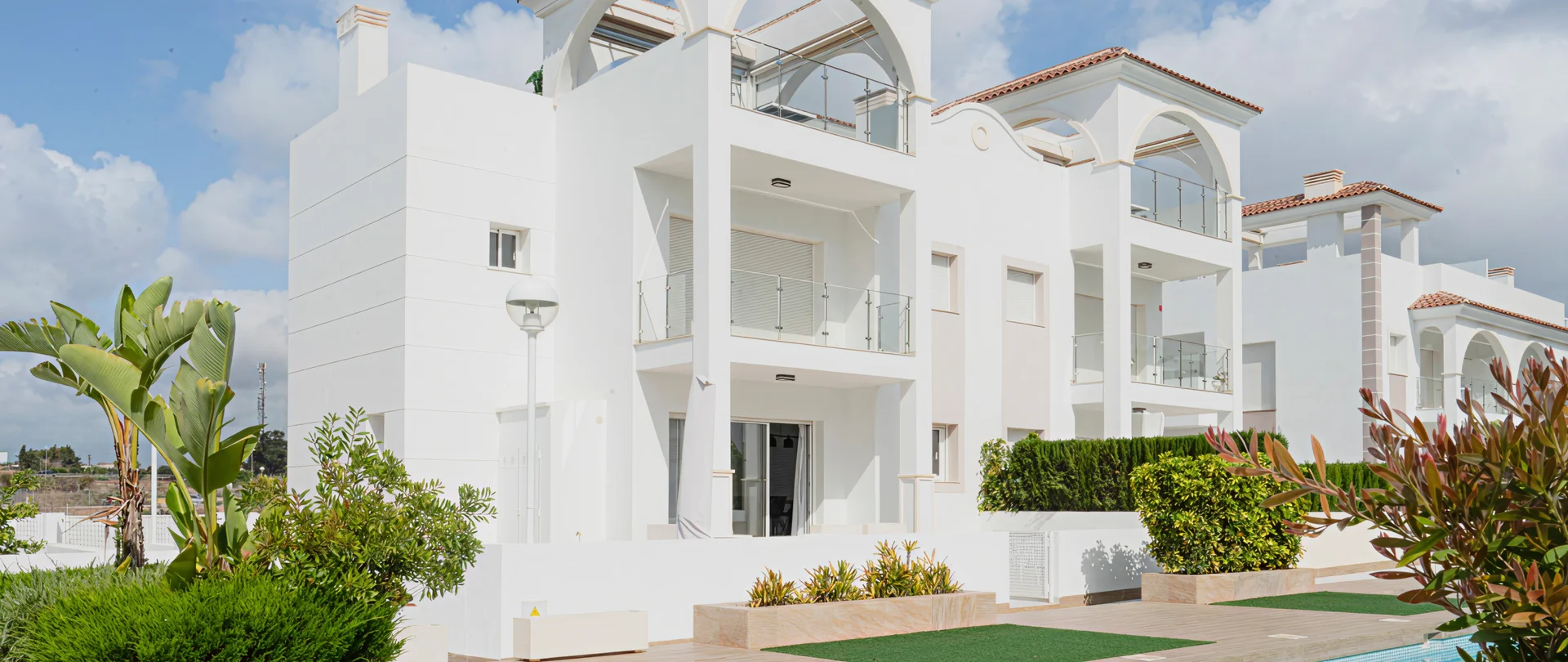A three-story white stucco apartment complex with glass balconies and a terracotta roof, surrounded by lush greenery.