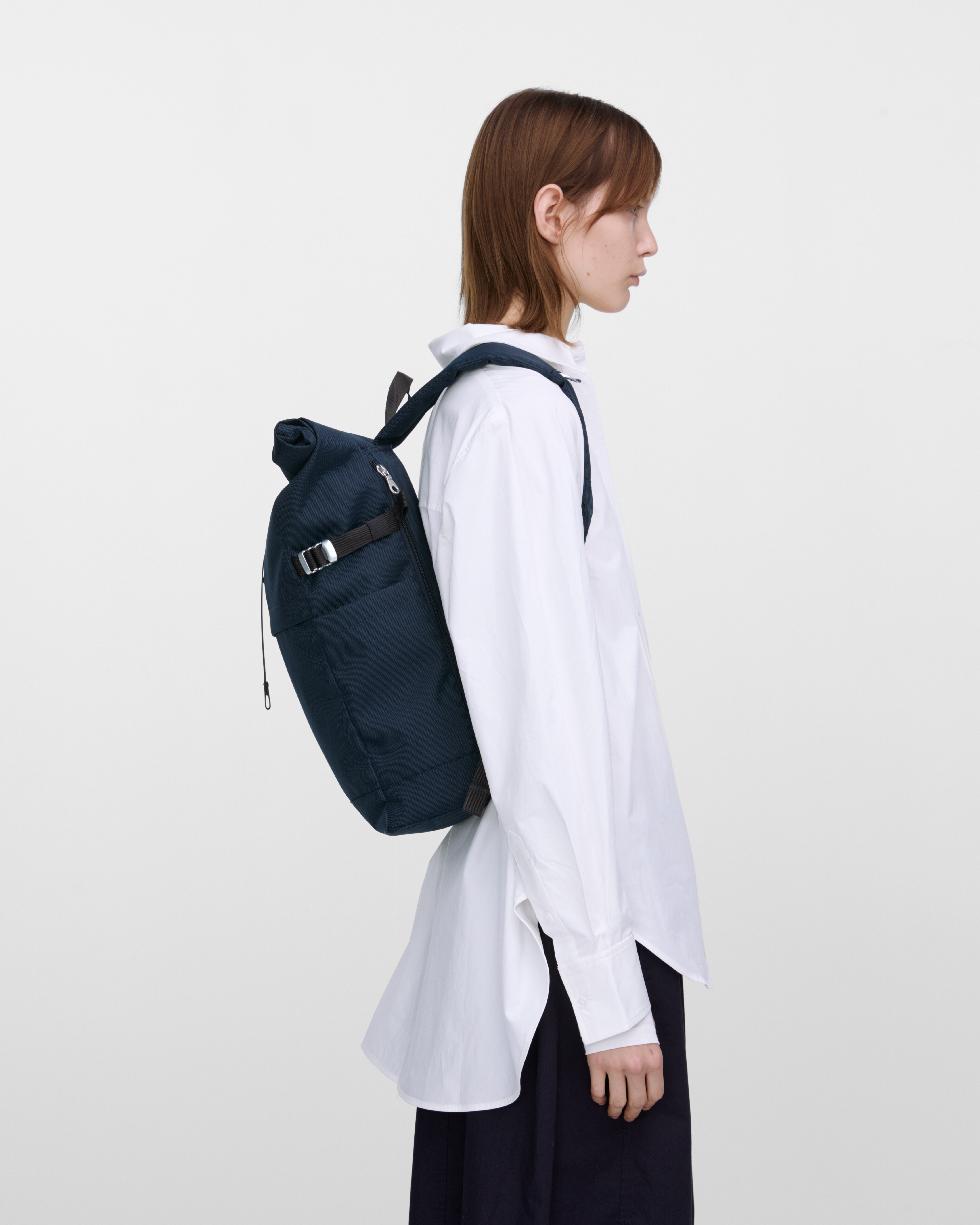 Female model carrying backpack Sandqvist ICON Rolltop Backpack M in dark blue, side view showing fit and size. 