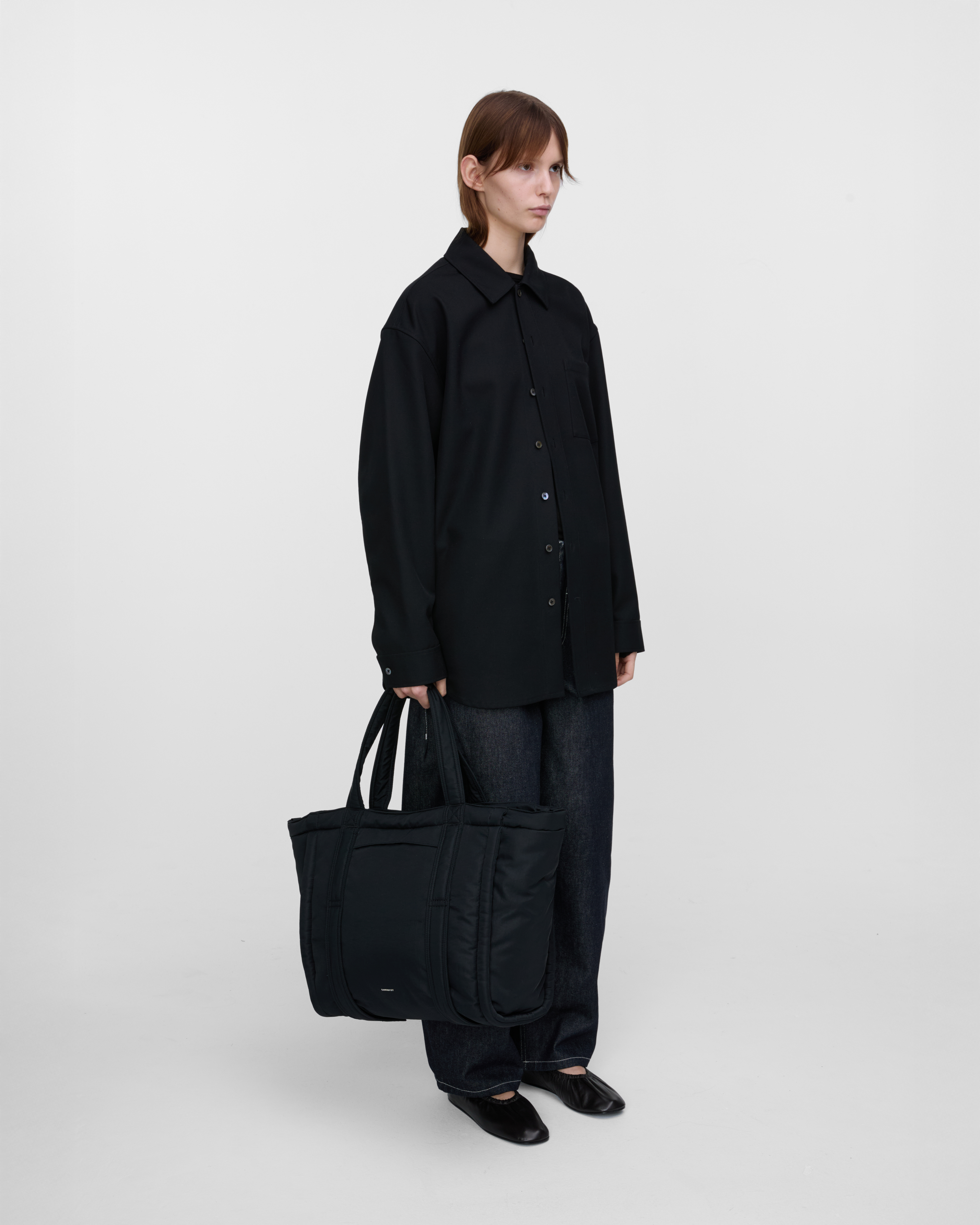 Female model holding Sandqvist Curve Tote Bag, black. Front view, holding the bag in one hand.