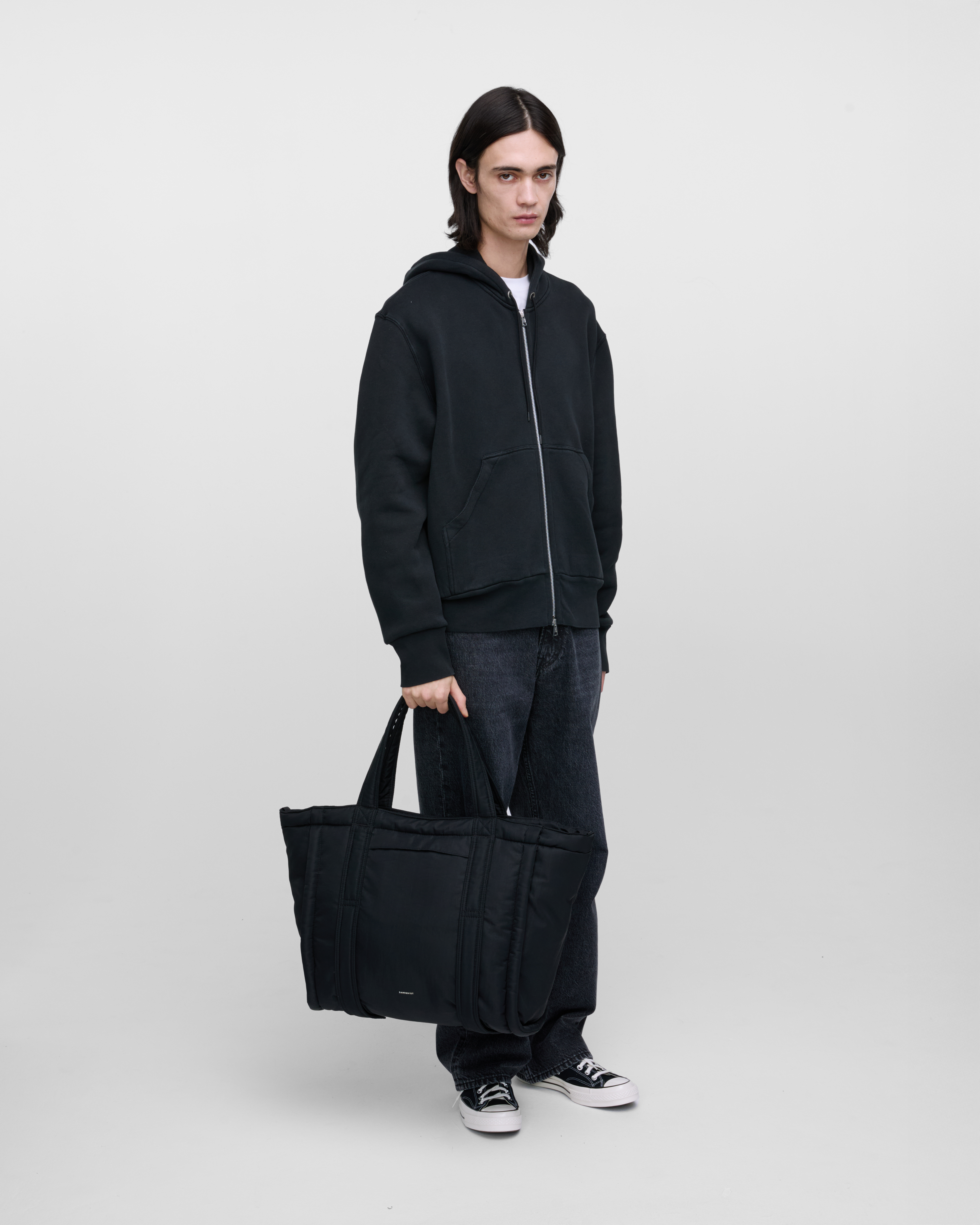 Male model holding Sandqvist Curve Tote Bag, black. Front view, holding the bag in one hand.